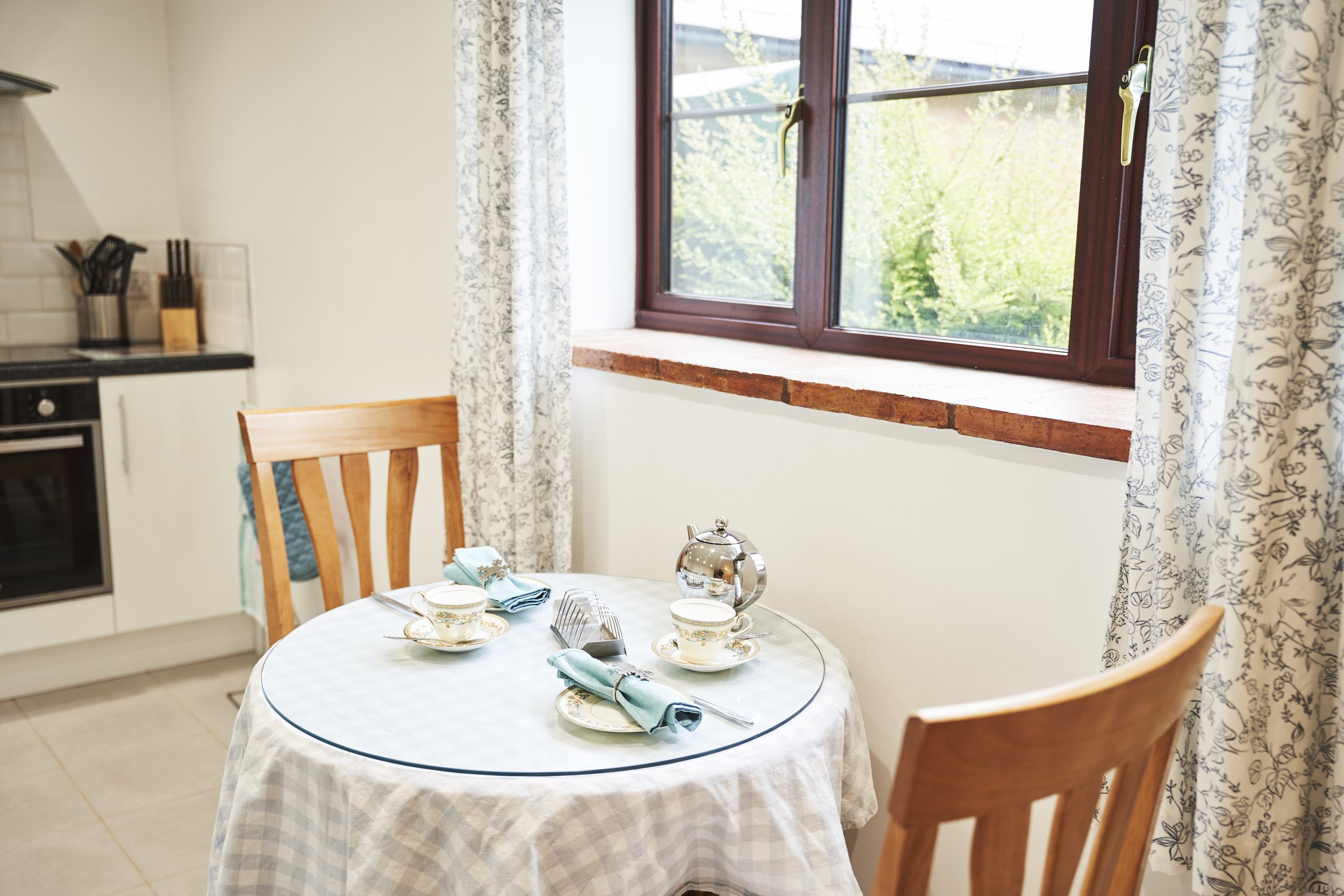 A cozy kitchen corner with a wooden dining table set for two, featuring floral teacups and teal napkins, near a window with floral curtains and a glimpse of greenery outside.