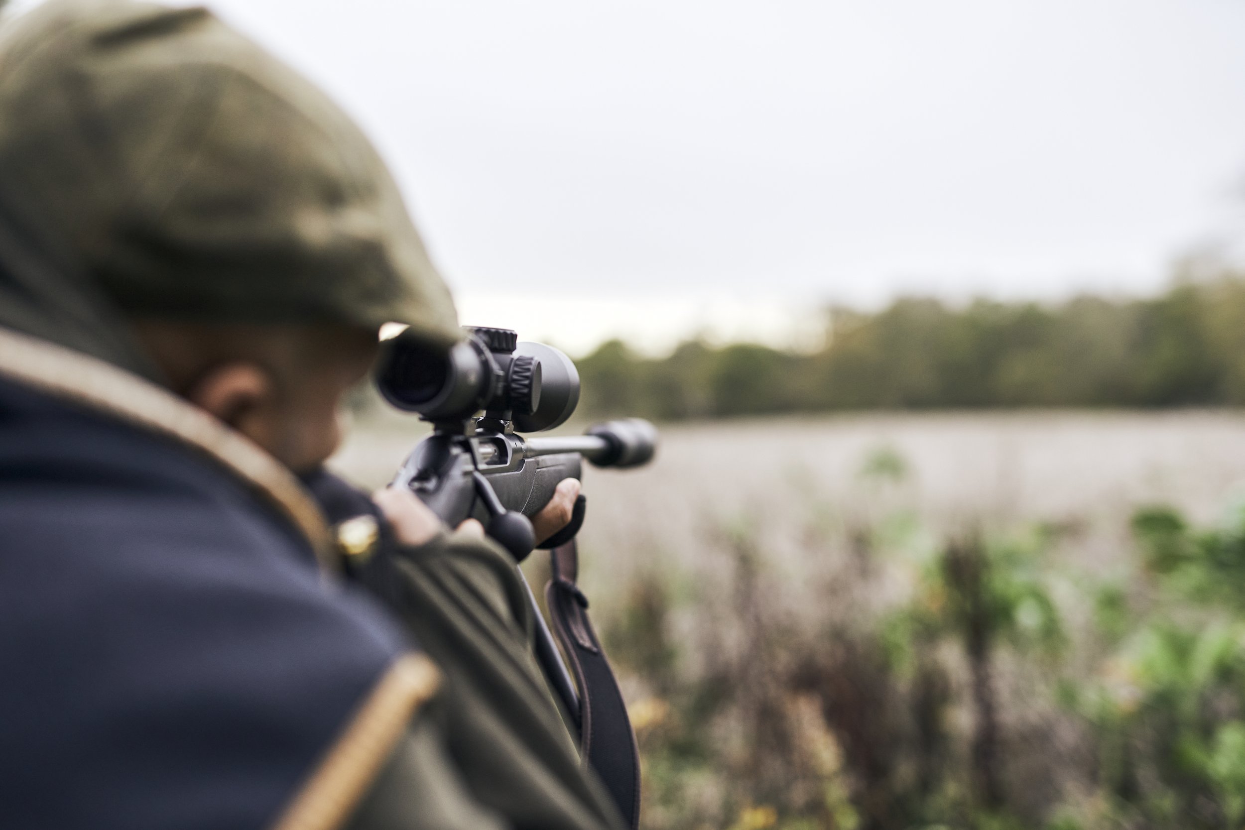 A person aiming a rifle with a scope in an outdoor field.