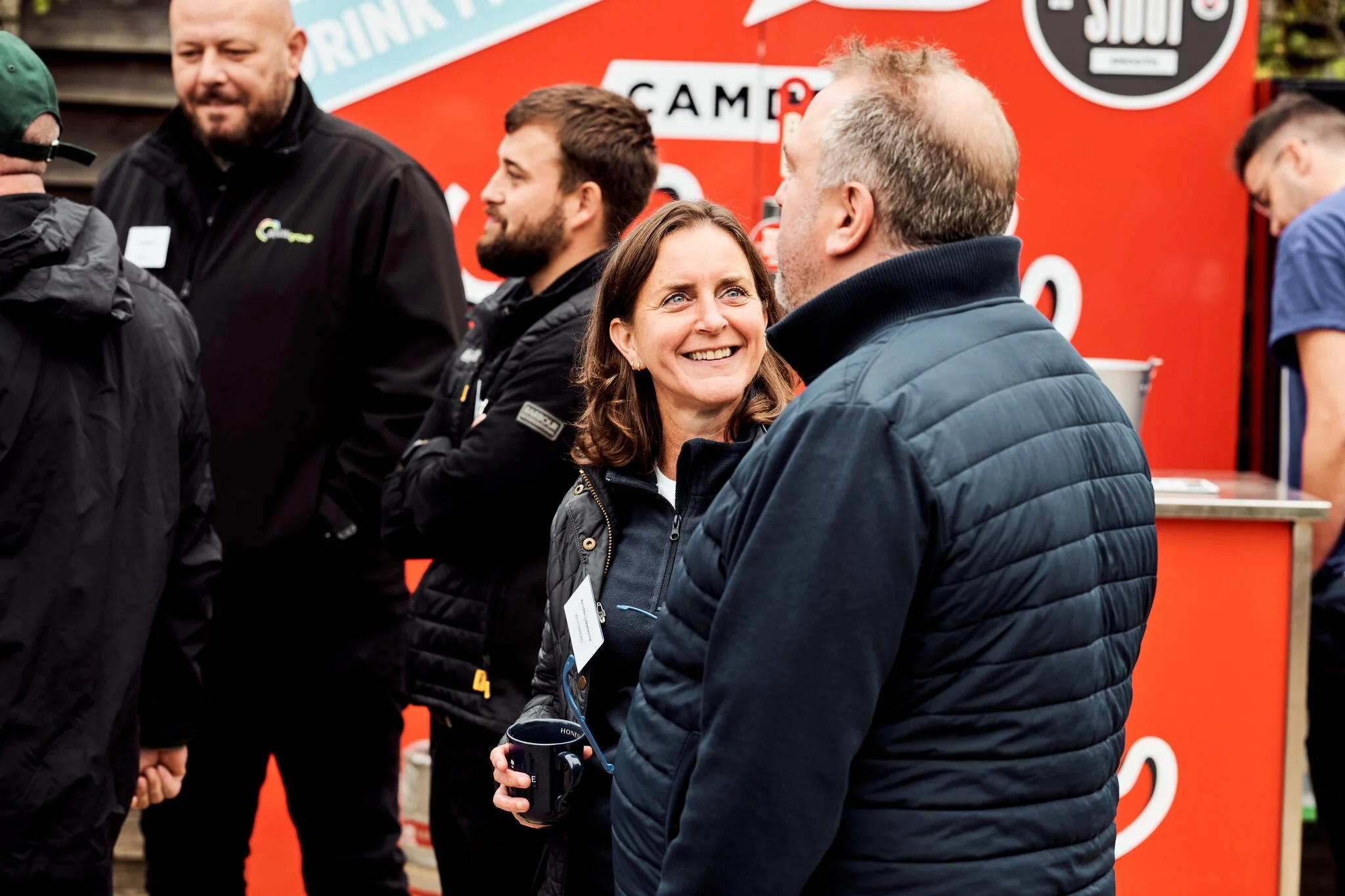 A group of people having conversations at an outdoor event, with a woman smiling and holding a mug, and a man talking to her, wearing a navy blue puffer jacket, with a red promotional booth in the background.