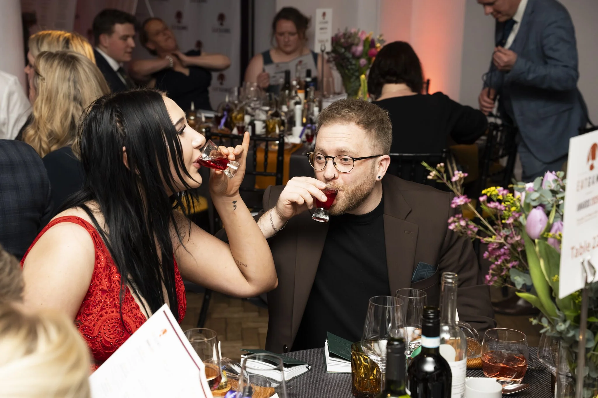 A woman in a red dress and a man in a black suit are sitting at a table drinking red wine at a social gathering or dinner event. There are flowers, bottles, and glasses on the table. Other people are seated around, engaging and looking at menus or co