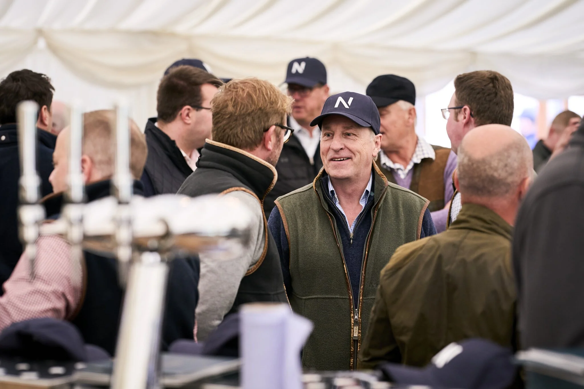 Group of men talking at an outdoor event under a white canopy tent, some wearing hats and jackets, with blurred objects in the foreground.