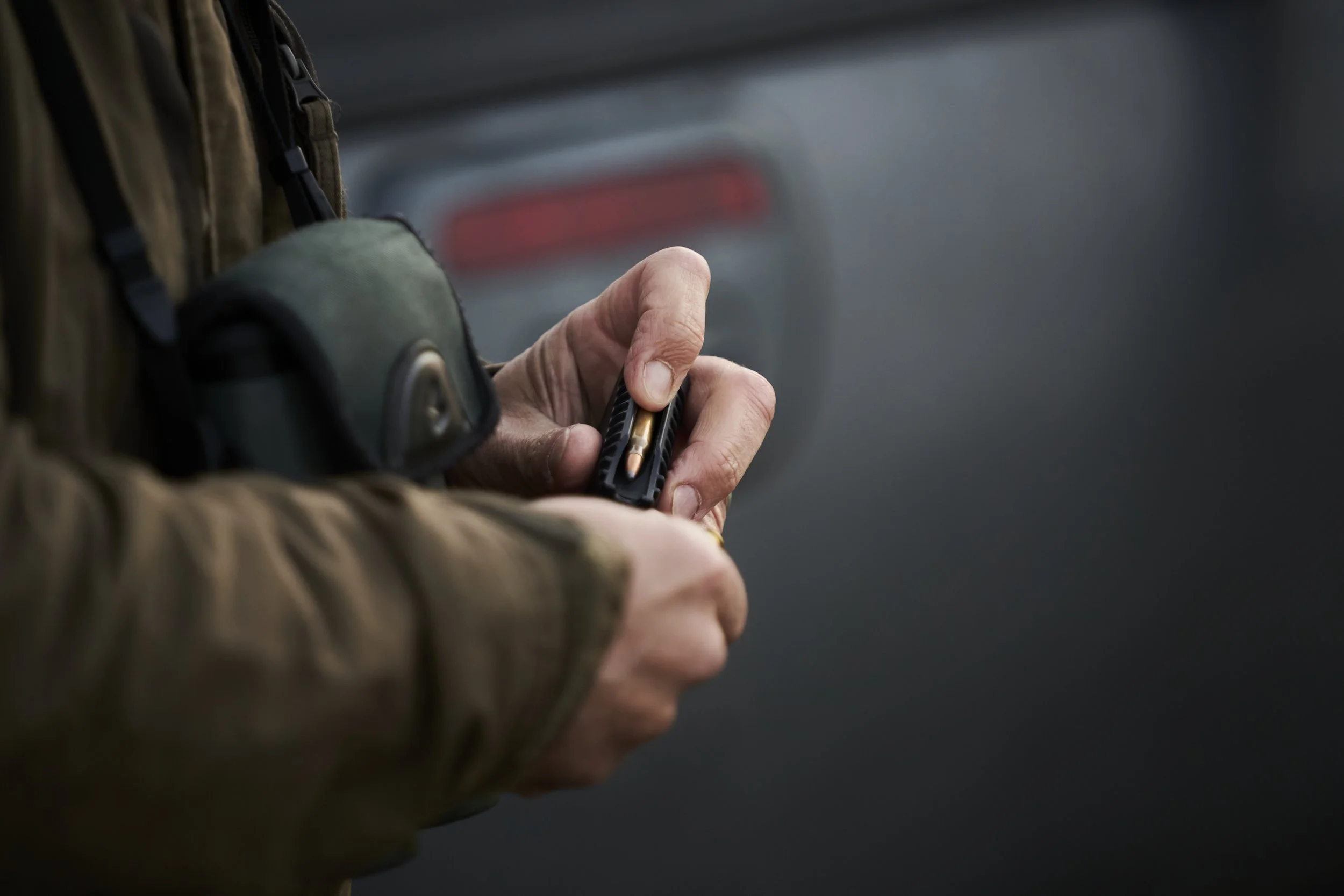 Close-up of a person in outdoor gear holding a handgun with one hand and inserting a bullet into the magazine with the other hand, with a blurred vehicle in the background.