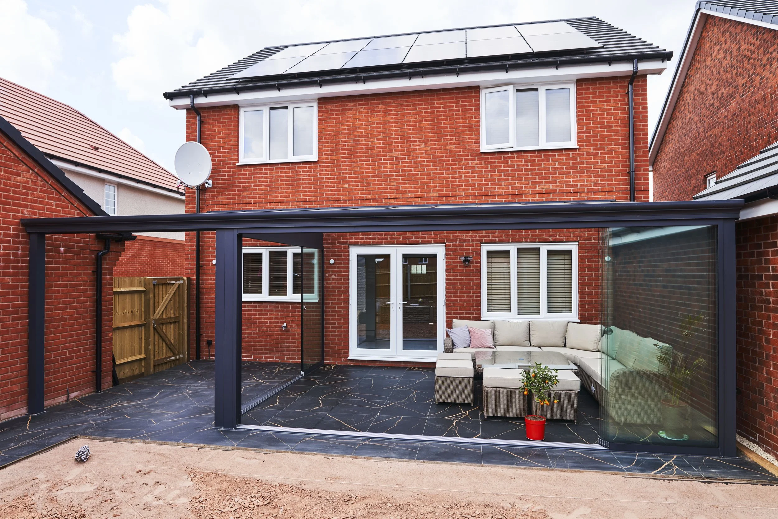 Backyard patio with black marble tiles, outdoor sofa with cushions, glass coffee table, and potted plant, enclosed by glass walls, attached to a red brick house with a patio door, and neighboring houses in the background.