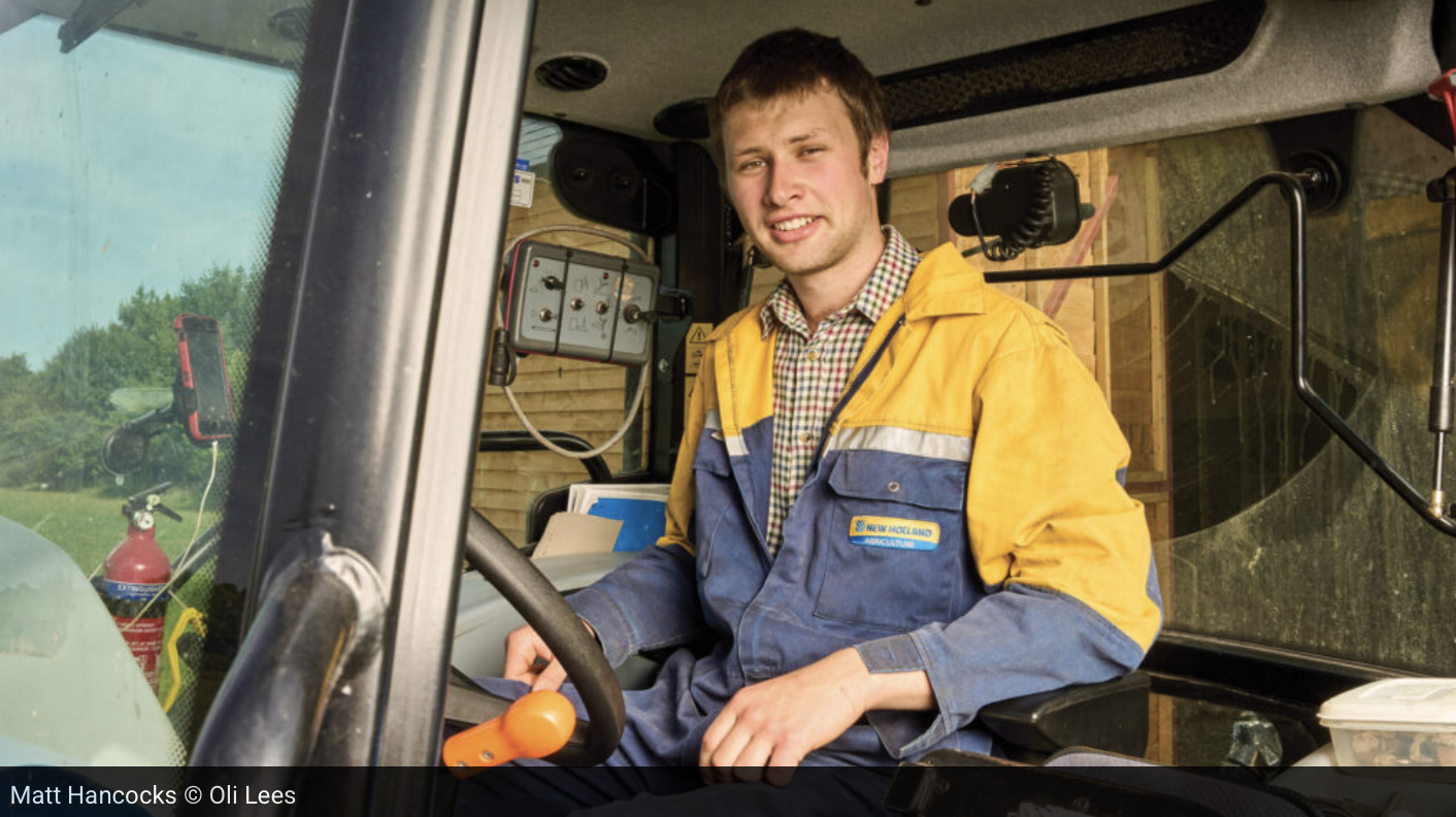 Young man in yellow and blue work jacket sitting in the driver’s seat of a construction vehicle, smiling.