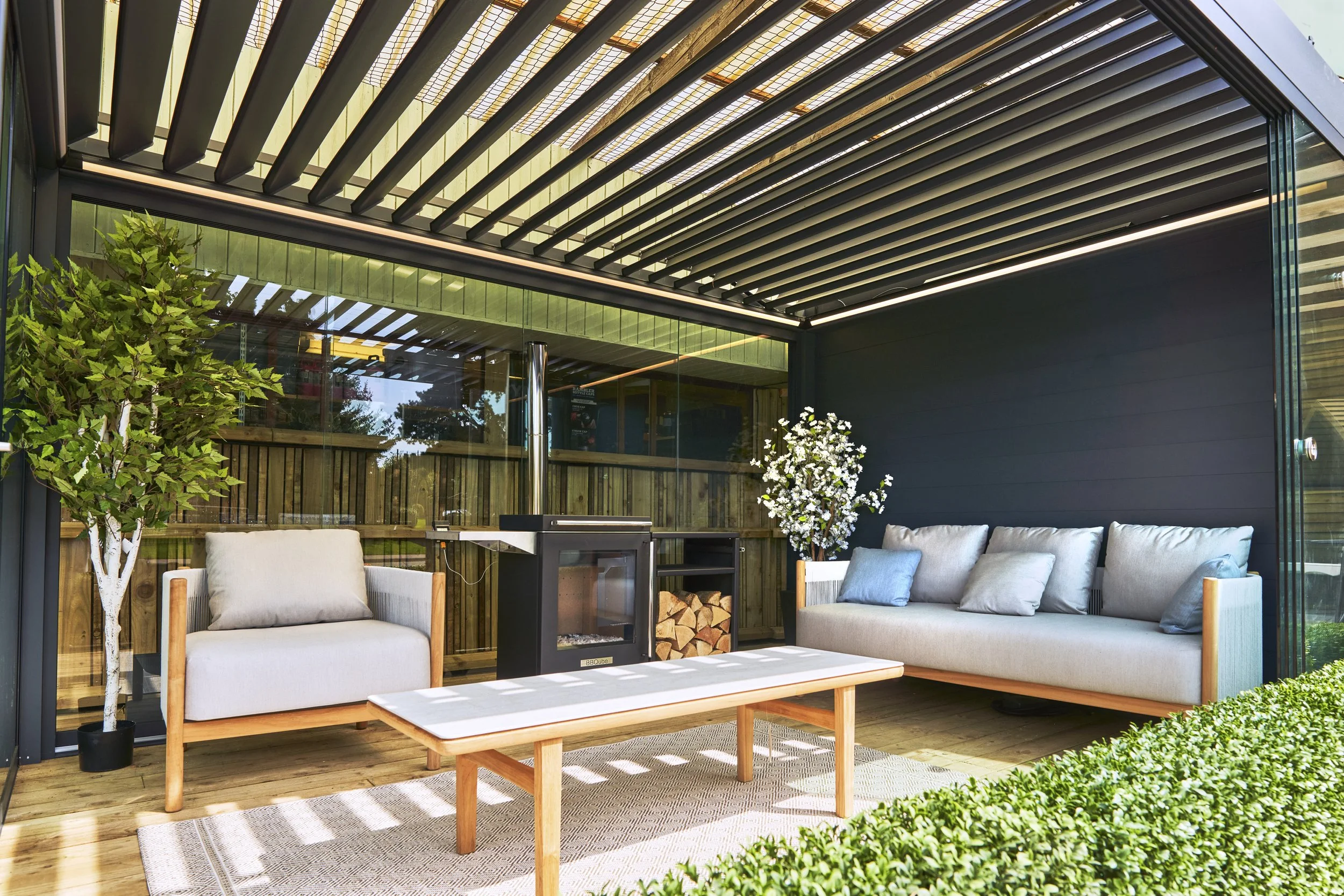 Outdoor patio with two modern sofas, a coffee table, a wood-burning stove, potted plants, and a green hedge, under a pergola with wooden slats.