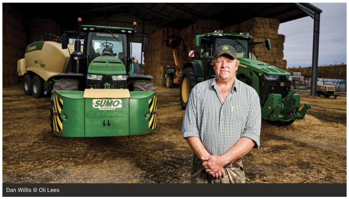A farmer standing in a barnyard with two green John Deere tractors and stacked hay bales in the background.