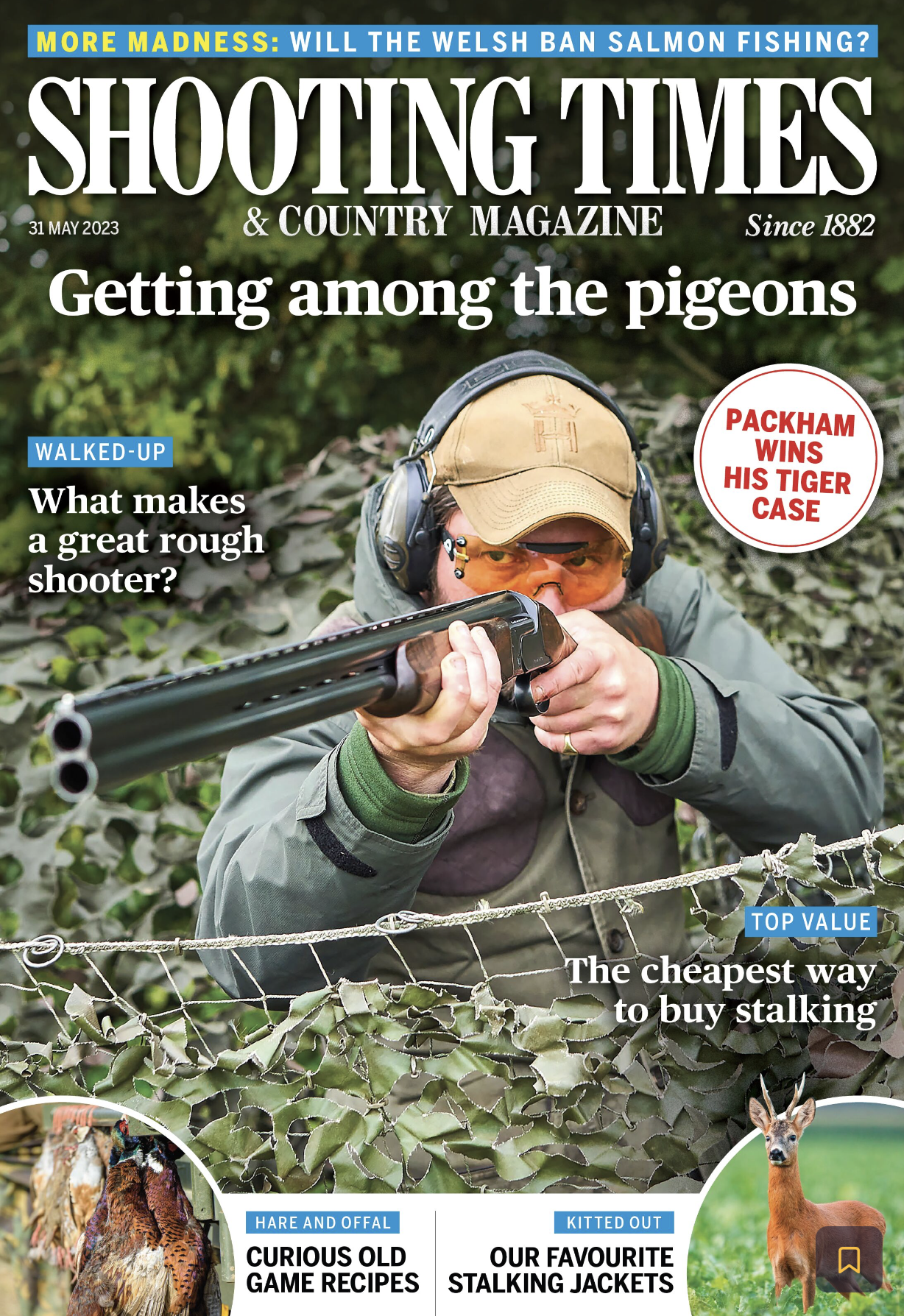A man wearing headphones and orange safety glasses aiming a shotgun through a camouflage net in a wooded area.