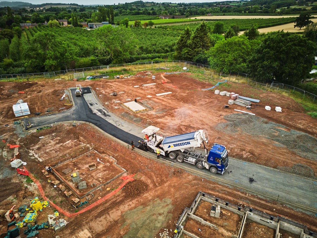 A construction site with a paved road curving through, surrounded by dirt and building materials. Construction workers and a cement mixer truck labeled 'BREEDON' are present, with greenery and farmland in the background.