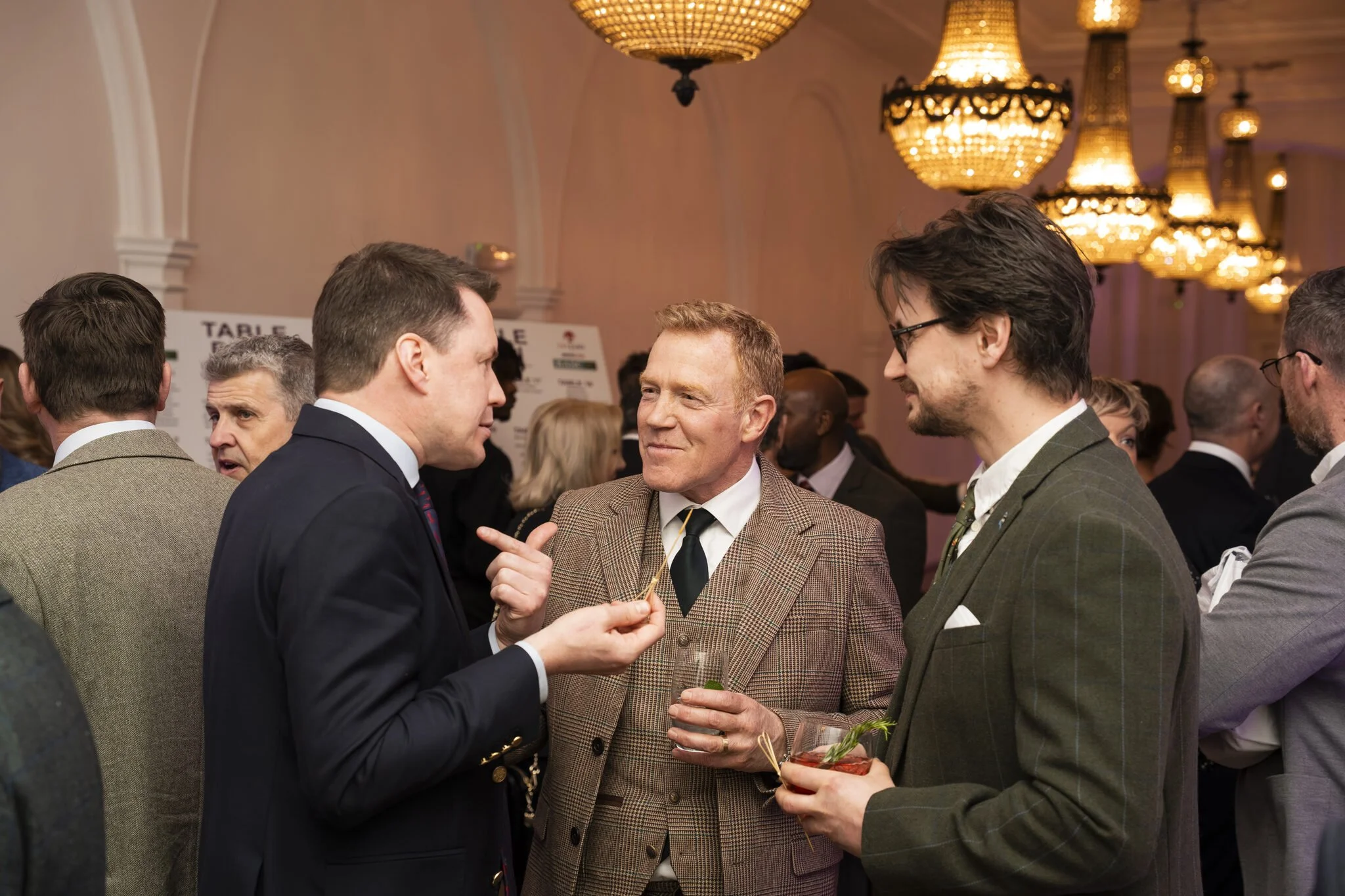 Group of men in suits conversing at an event with chandelier lighting in a banquet hall.