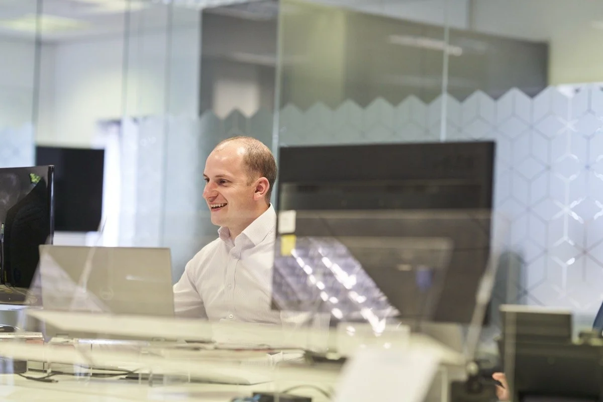 A man working at his desk in an office, smiling while looking at his computer monitor.
