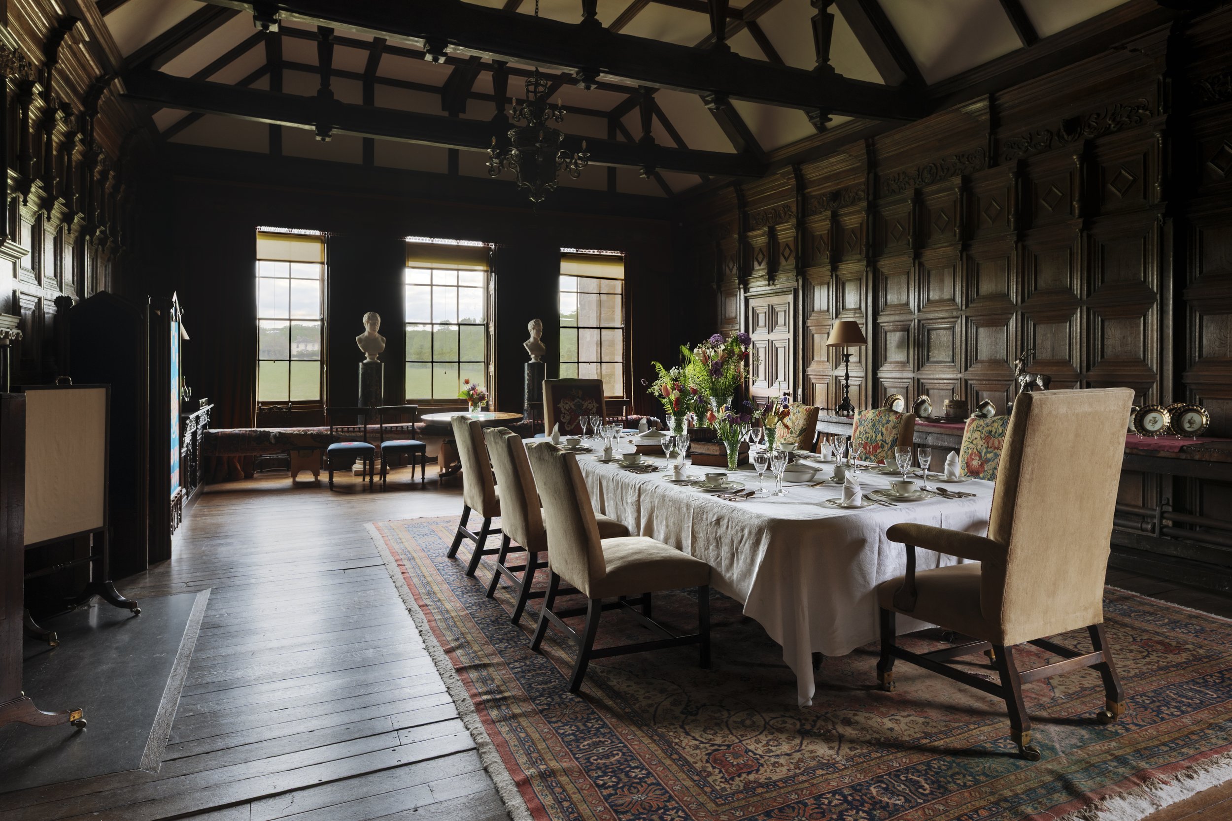A historic dining room with a long table set for a meal, surrounded by upholstered chairs, large windows, and dark wood-paneled walls with intricate carvings.