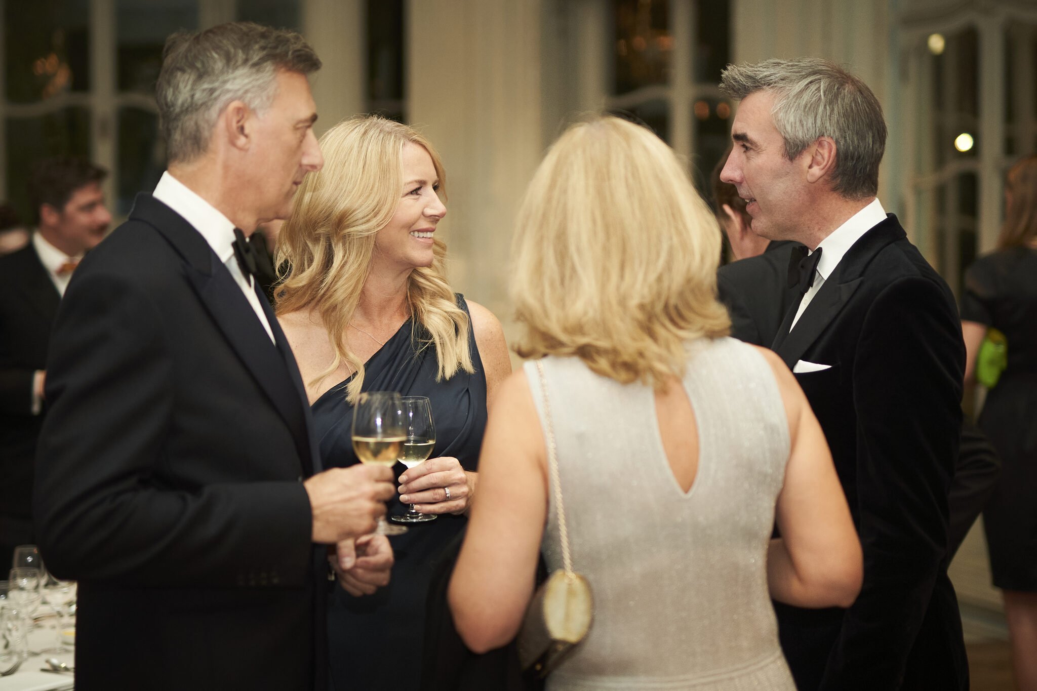 People dressed in formal attire at a social gathering, smiling and holding glasses of champagne, engaging in conversation indoors with large windows in the background.