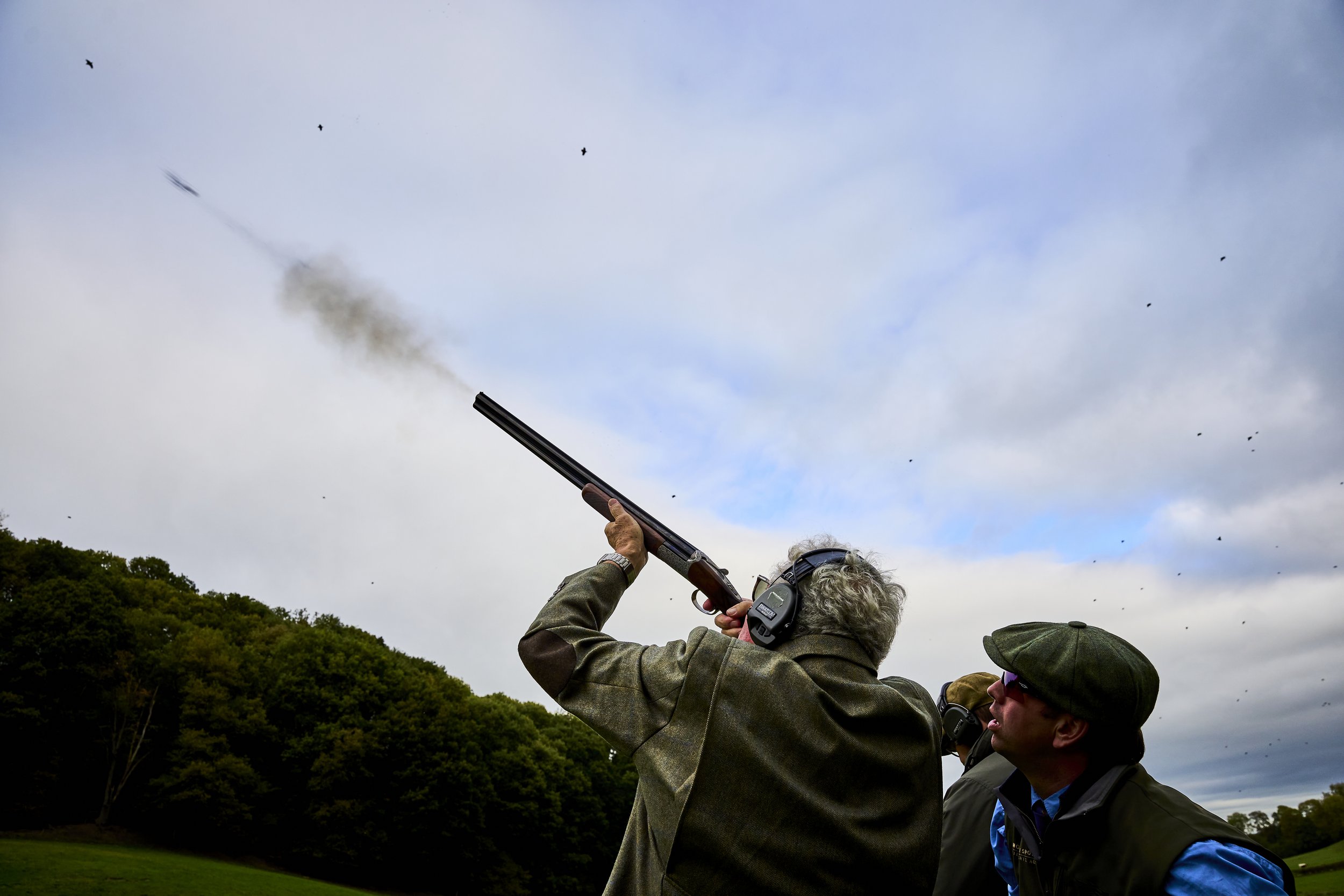 A man with curly gray hair wearing hearing protection and a jacket is aiming a shotgun, which is firing and releasing smoke, at a shooting event outdoors. A person in sunglasses and a beret is watching him.