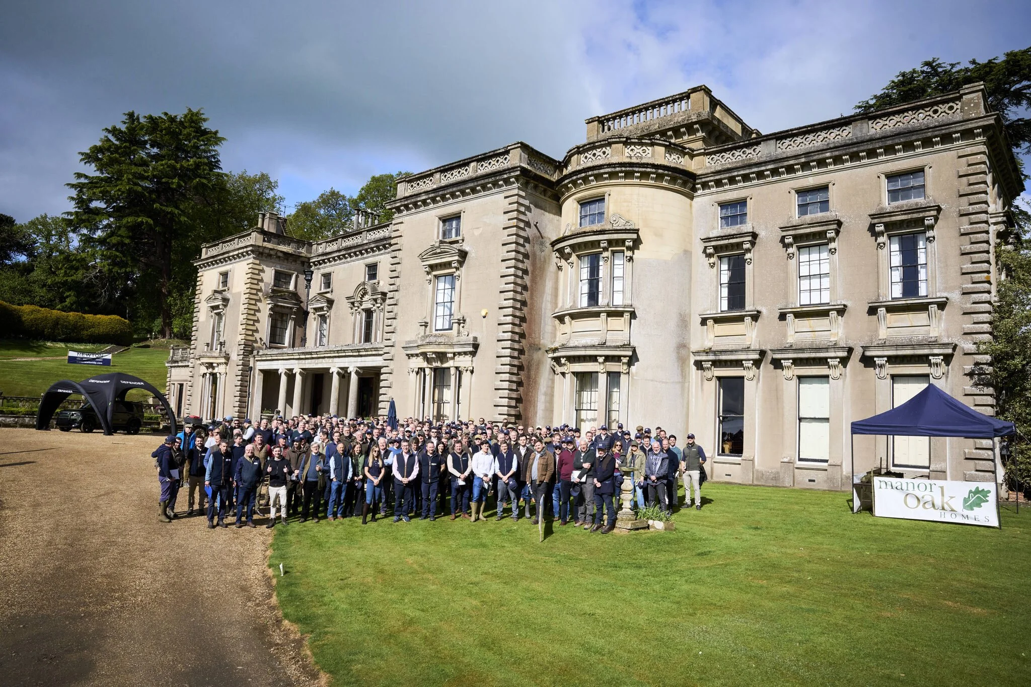 Large group of people gathered in front of a historic building with classical architecture, greenery, and partly cloudy sky.