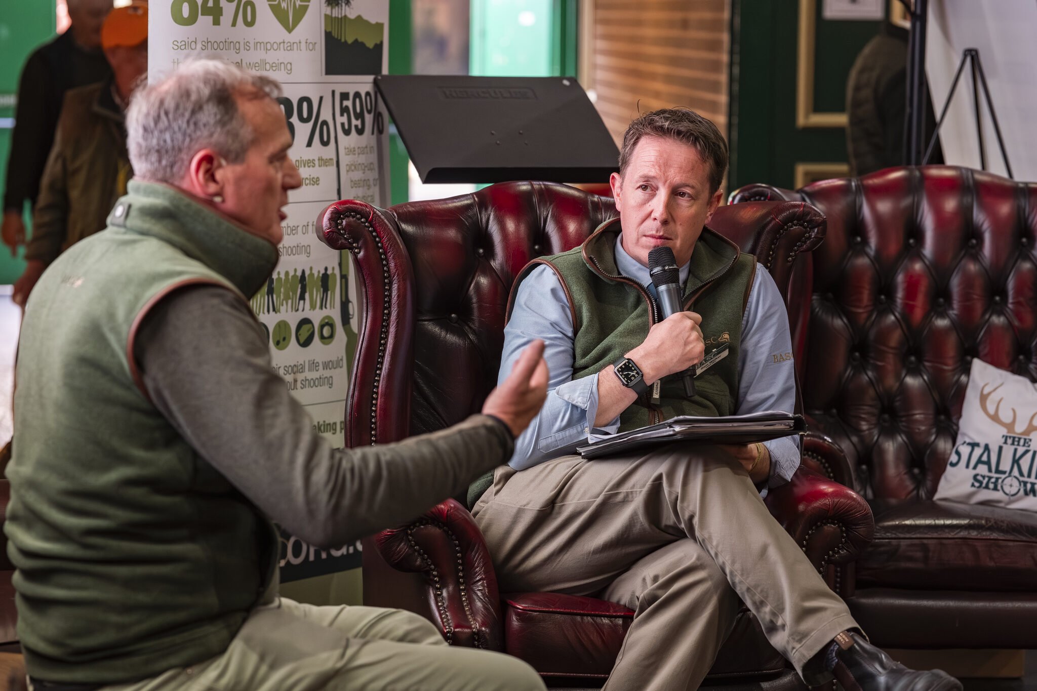 Two men sitting on a leather couch, participating in a discussion with one holding a microphone. A poster with statistics and green foliage can be seen in the background.