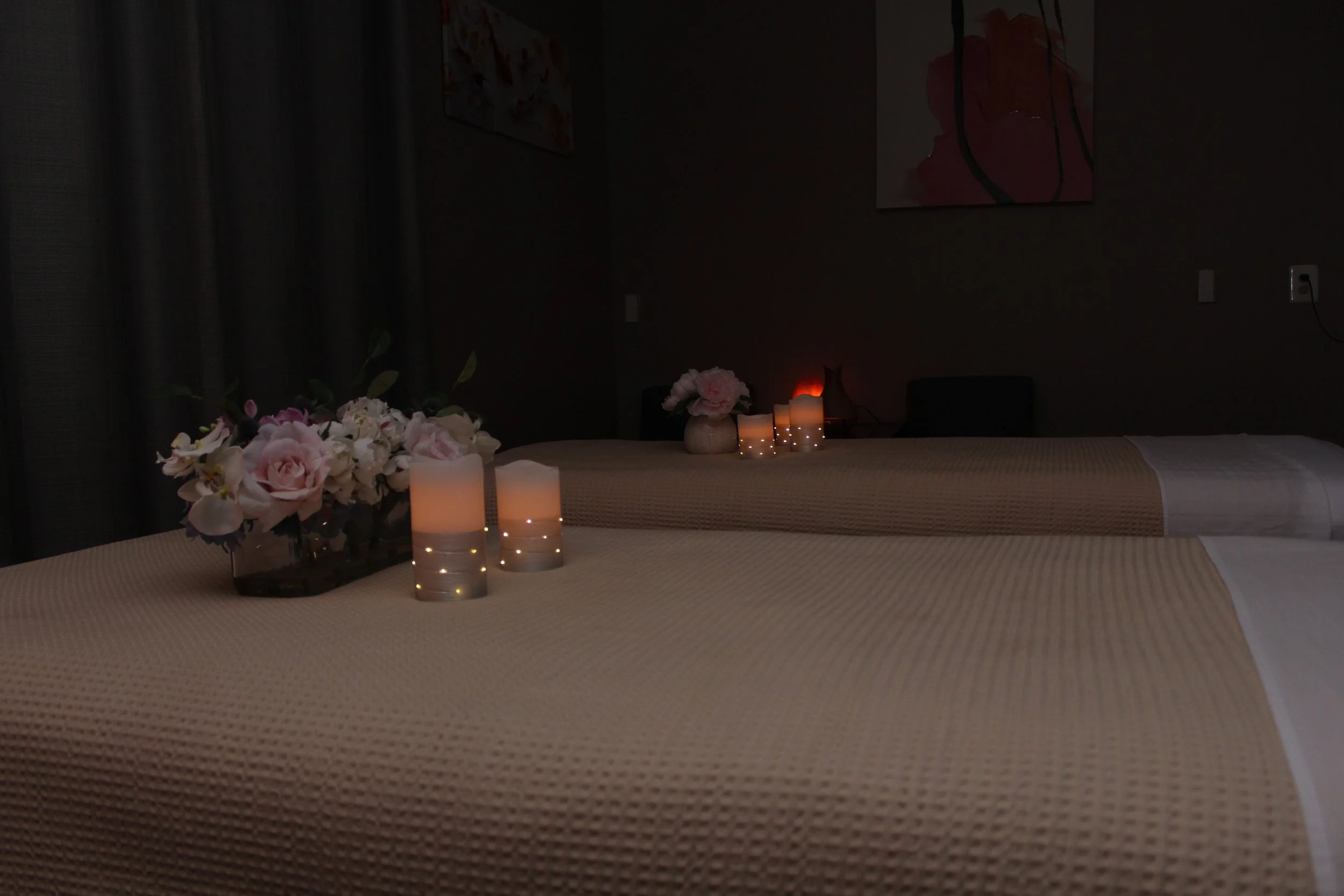 Two beds with beige bedding, decorated with floral arrangements and lit candles, in a dimly lit room.