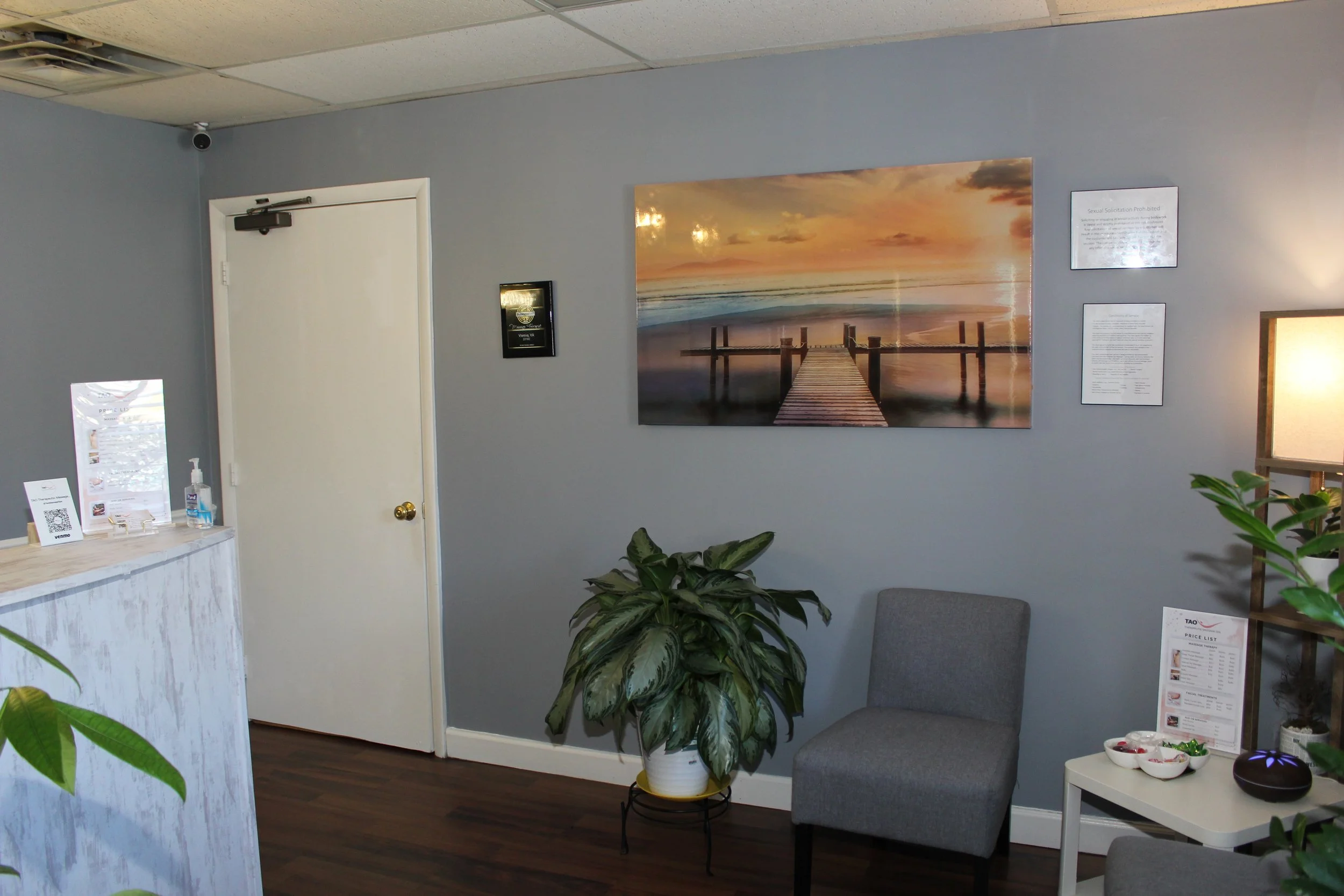 Interior view of a waiting room with gray walls, a gray chair, a large wall art of a sunset over a dock, and a small table with bowls and a plant.