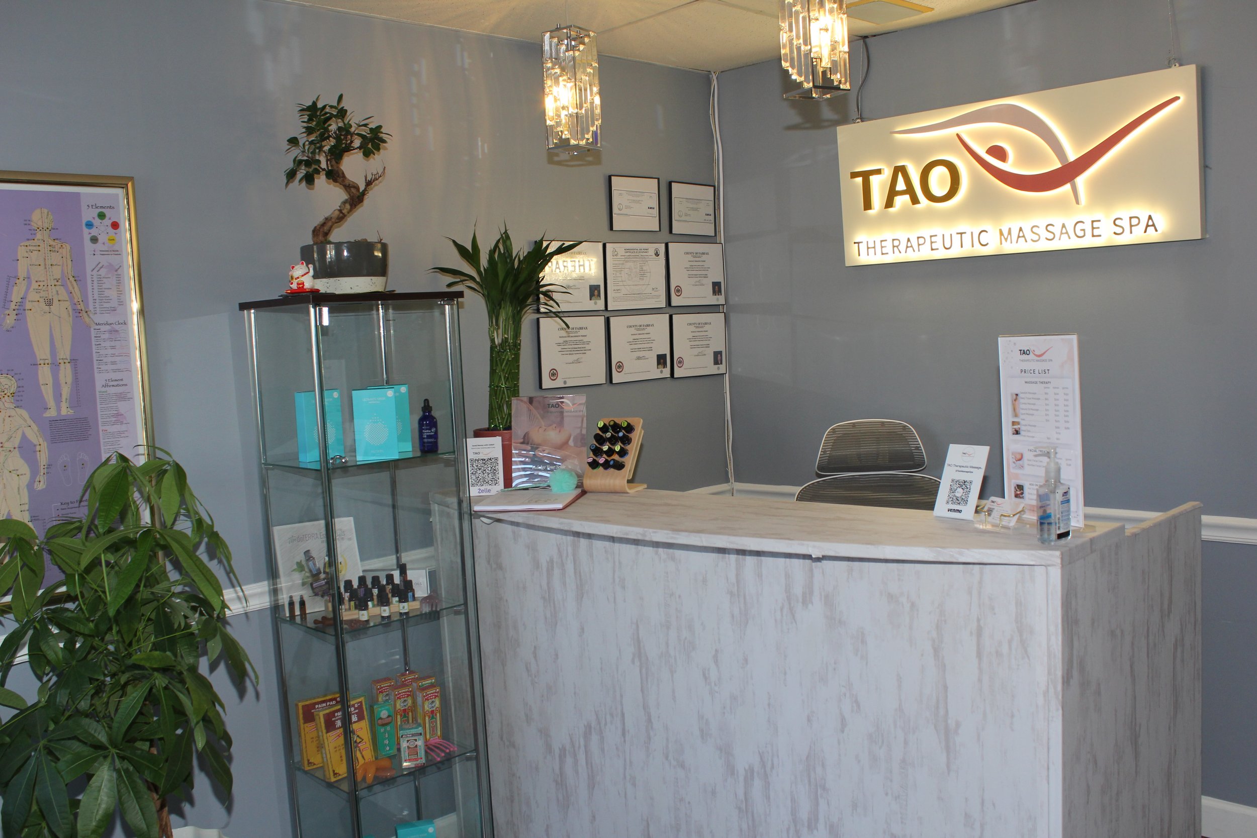 Reception area of a massage spa with a sign that says TAO Therapeutic Massage Spa. There is a reception desk, a glass display case with massage oils and products, framed certificates on the wall, and potted plants. A QR code, sanitizer, and price list are on the counter.