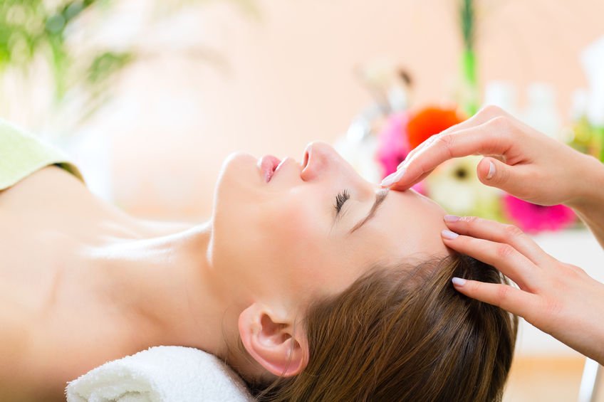 A woman is lying on her back with her eyes closed, receiving a facial treatment from a practitioner who is applying a product to her forehead. The setting appears to be a spa or skincare clinic with a blurred background including flowers.