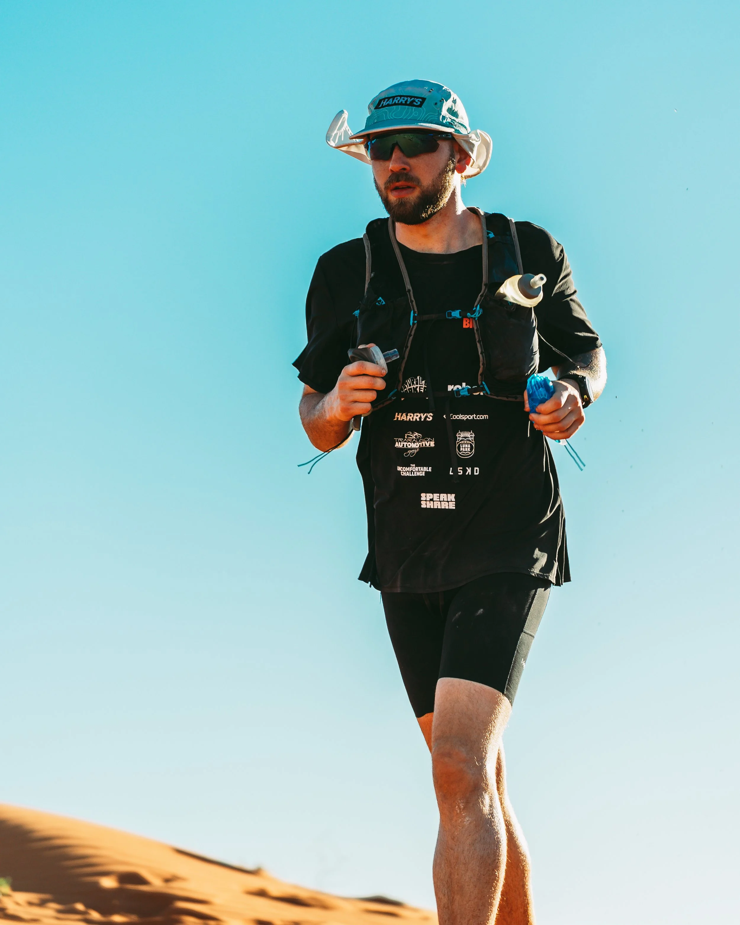 A man running in a desert landscape with sand dunes and clear blue sky, wearing a hat, sunglasses, and athletic gear.