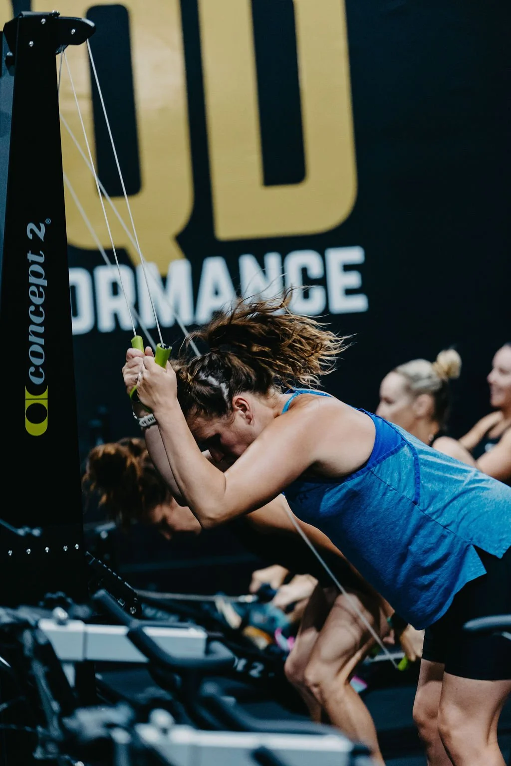 Women participating in an indoor HYROX class, wearing athletic clothing, with a large sign reading 'Performance' in the background.