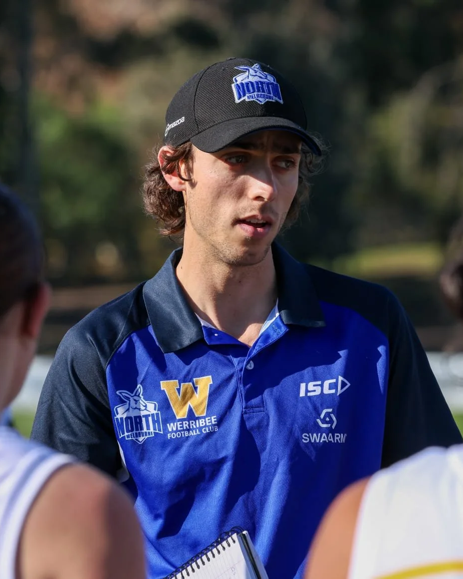 A young man with brown hair wearing a black cap and a blue sports shirt with logos, speaking to two athletes during an outdoor sports event.