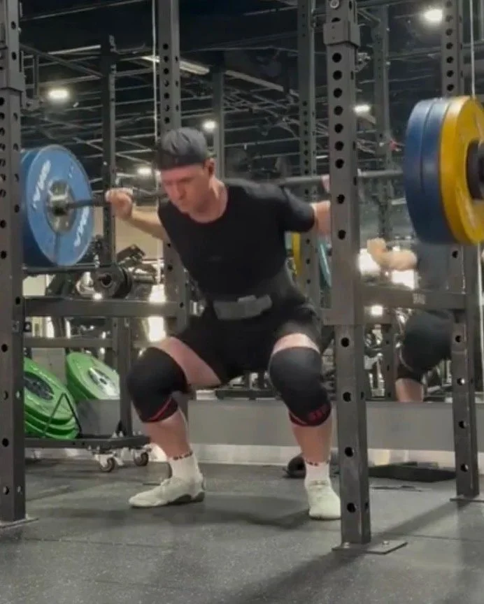 A man in gym gear performing a squat exercise with a weighted barbell in a gym.
