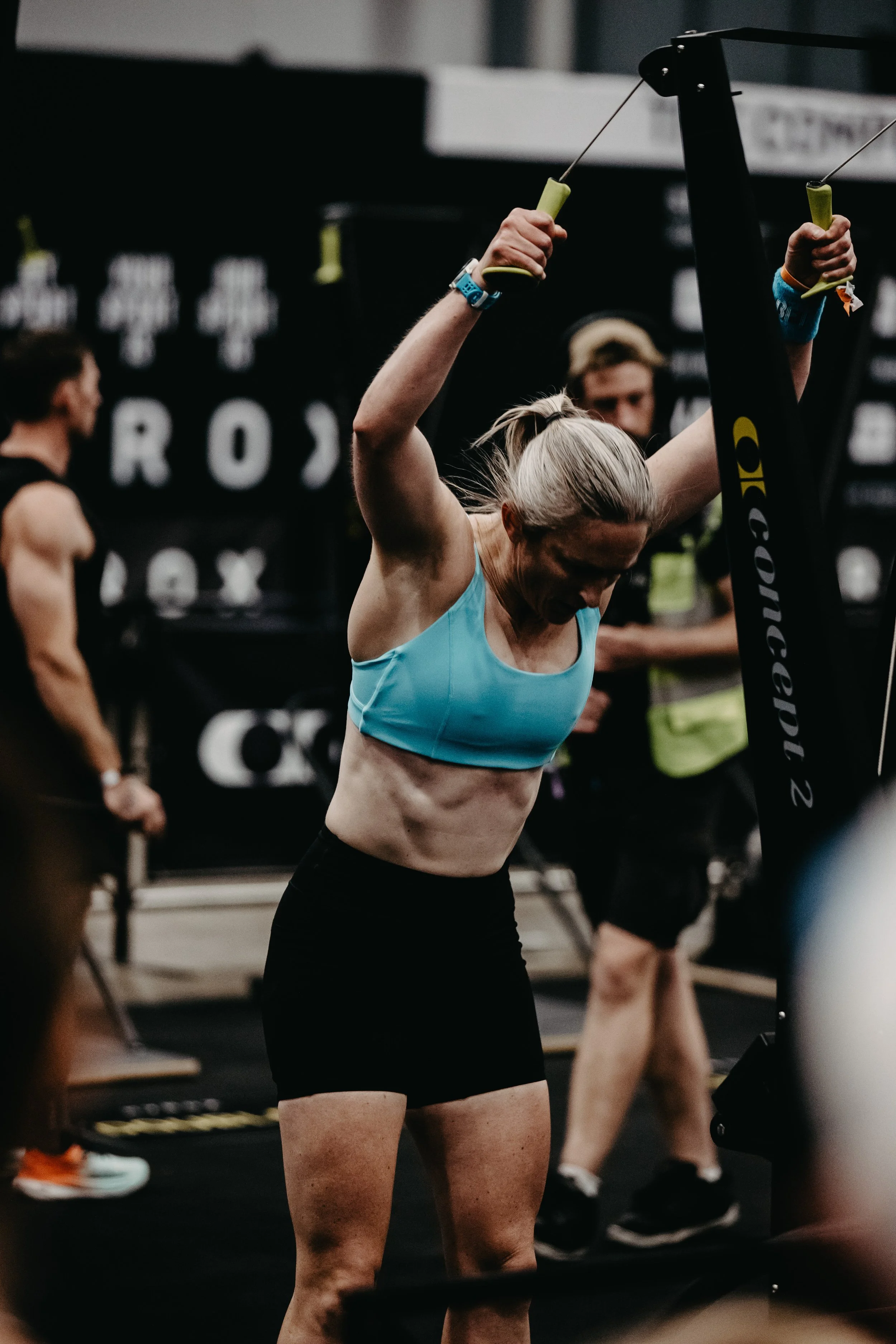 A woman with blonde hair in a ponytail wearing a light blue sports bra and black shorts, on the ski erg in a hyrox race