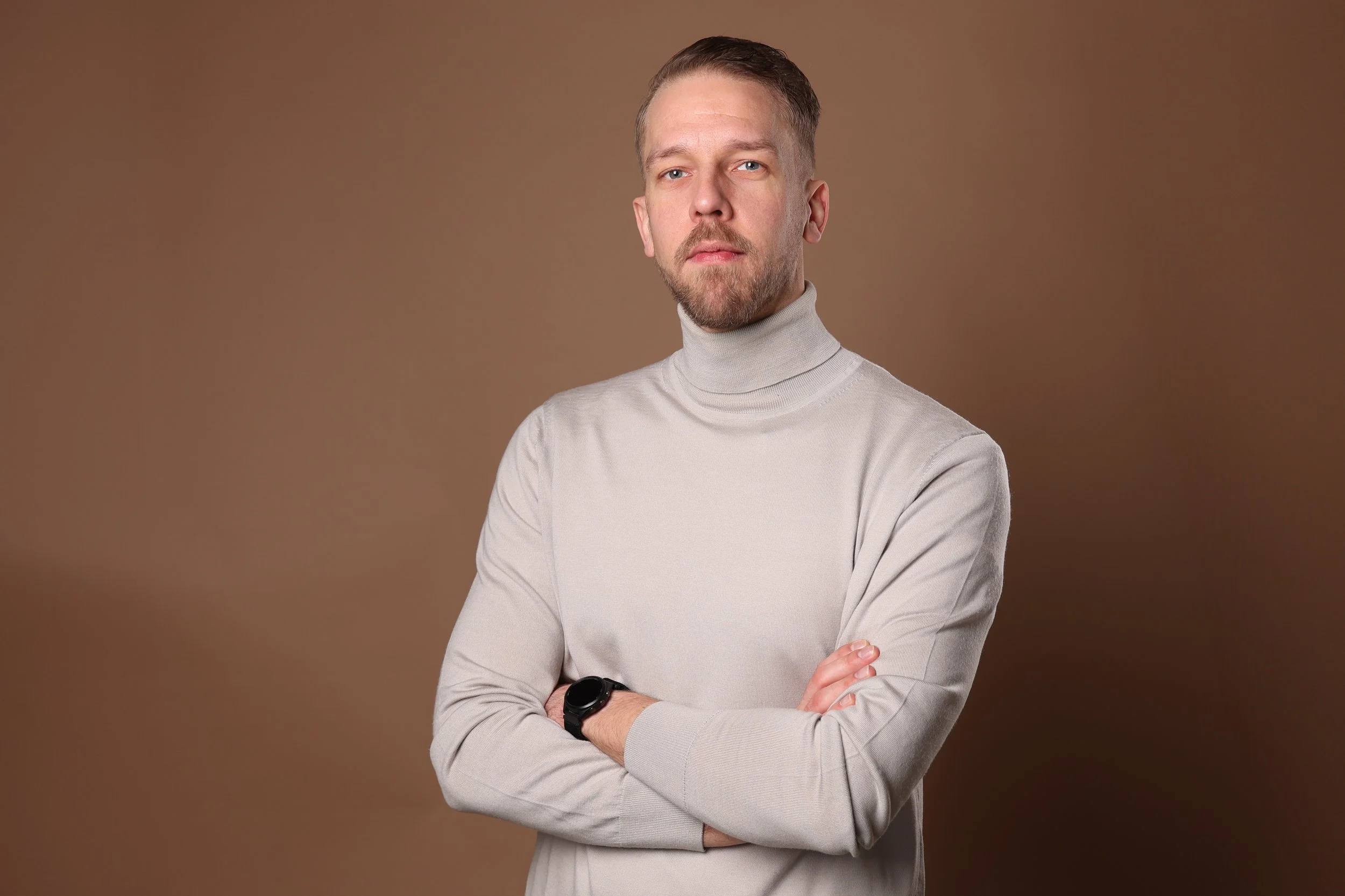 A young man with short hair and a beard, wearing a light grey turtleneck sweater, standing with arms crossed against a brown background.