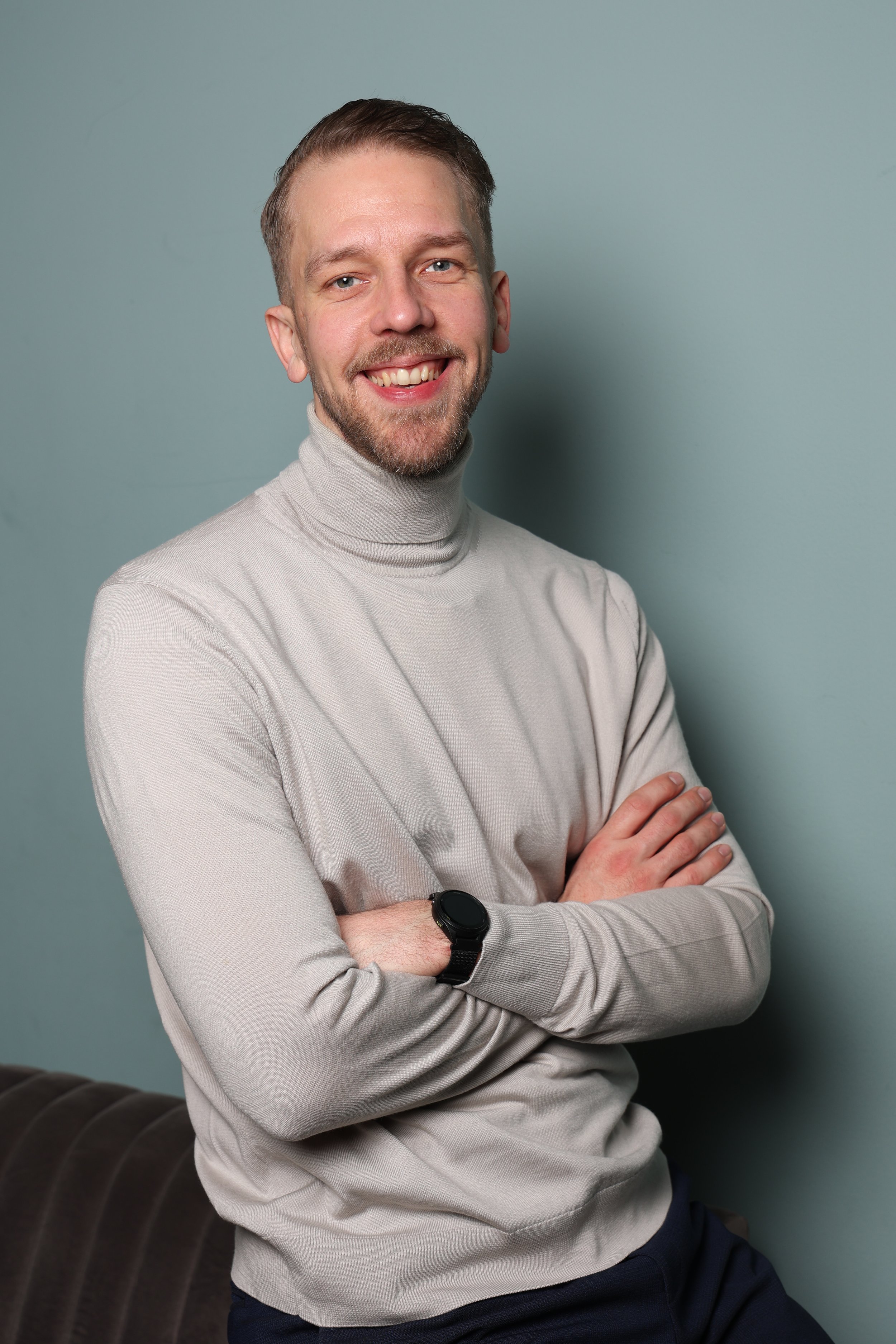A smiling man with light brown hair and a beard, wearing a light gray turtleneck sweater and a smartwatch, standing with arms crossed against a plain background.