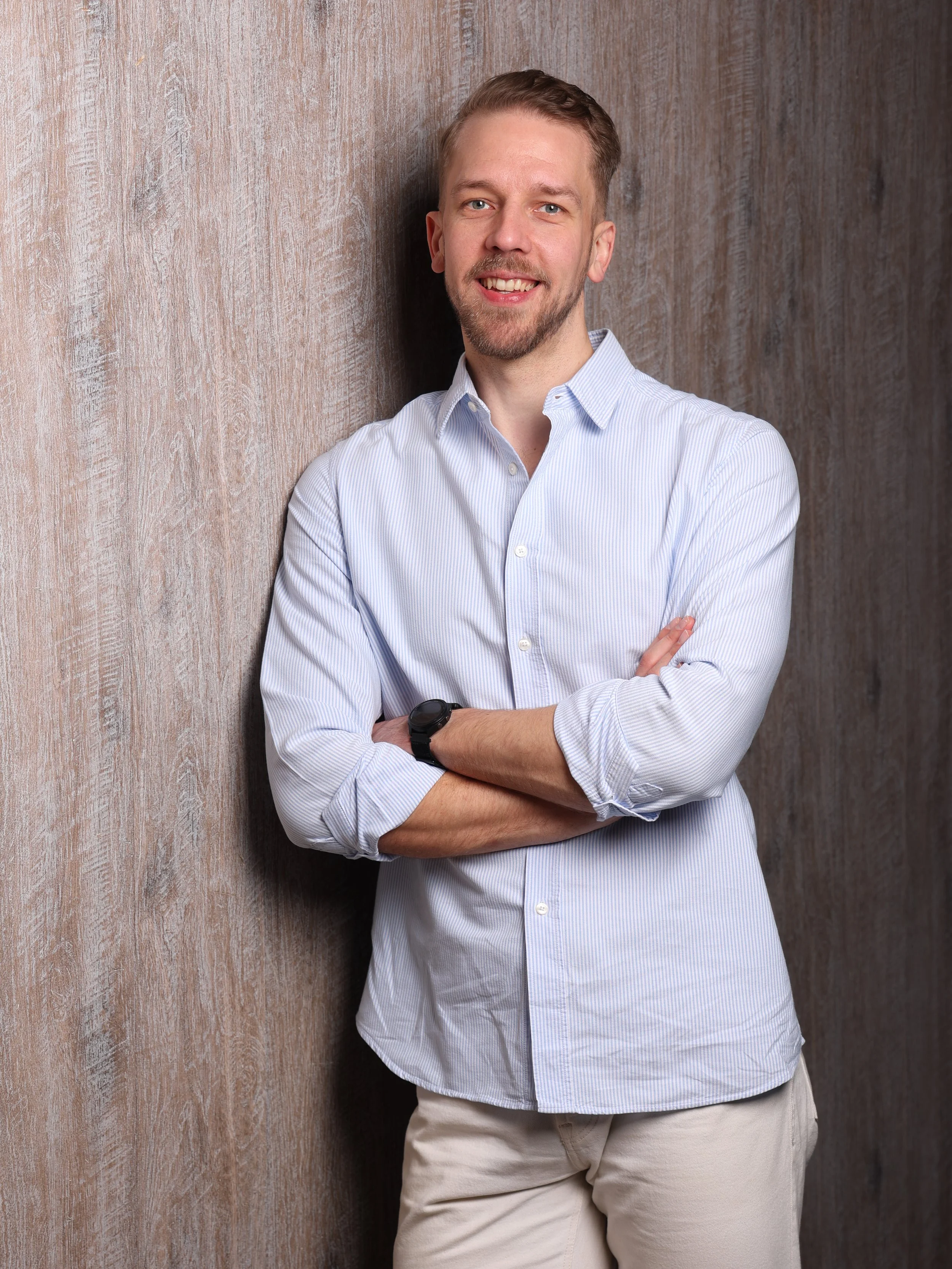 A smiling man leaning against a wooden wall, wearing a light blue button-up shirt with the sleeves rolled up and beige pants.