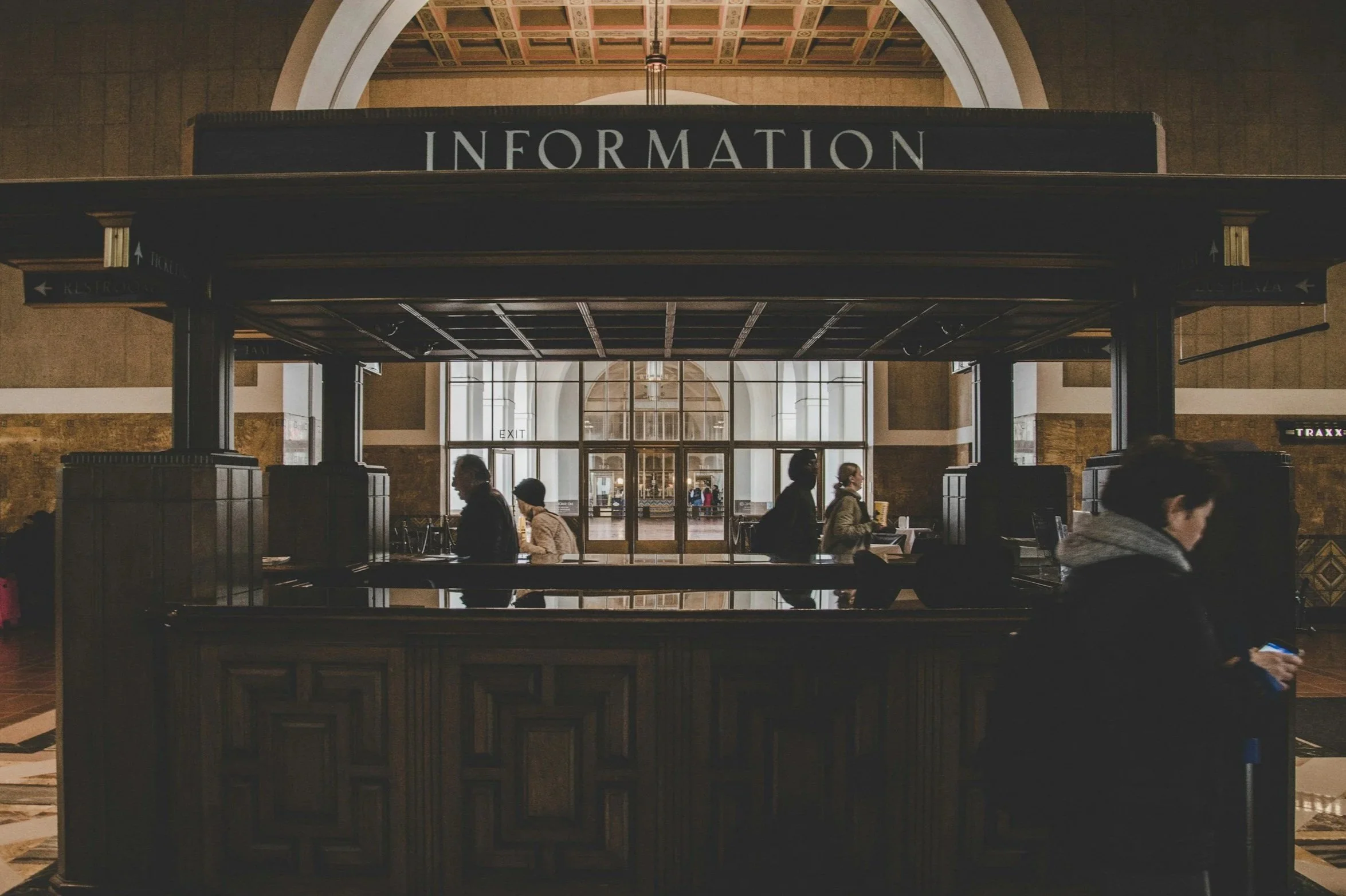 An information booth inside an airport with a black sign labeled 'INFORMATION' and several people walking past or waiting at the counter, with large glass doors and arched ceiling in the background.