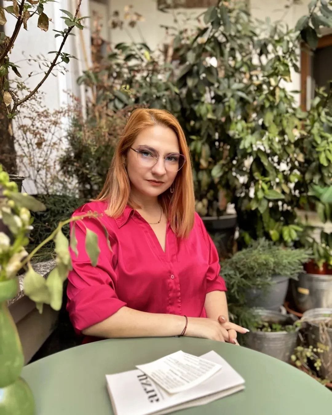 A woman with red hair, glasses, and a pink blouse sitting at a green table surrounded by potted plants and greenery.
