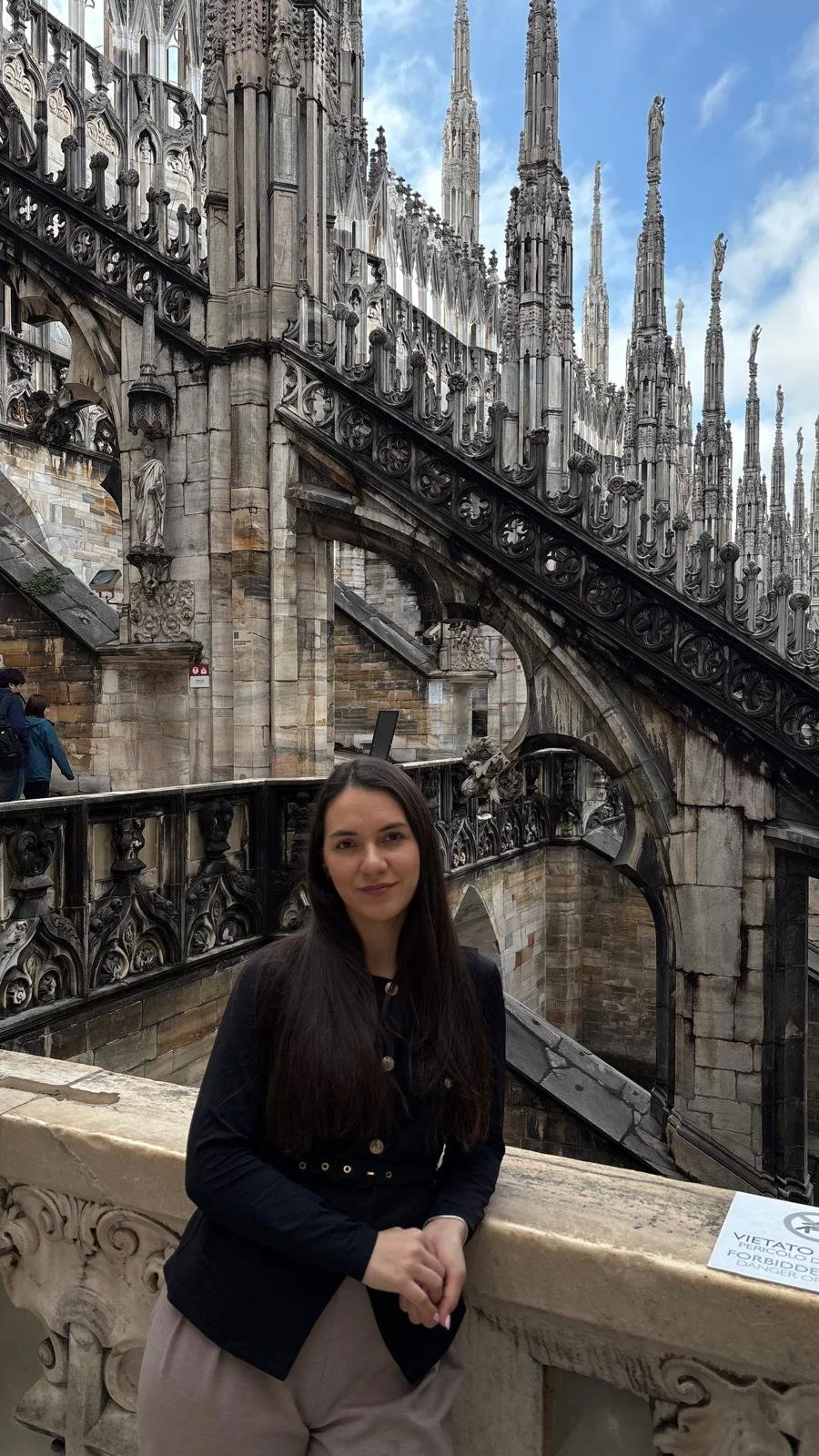 A young woman with long dark hair standing on a stone balcony at the top of a Gothic cathedral, with intricate stonework and spires behind her and a partly cloudy sky above.
