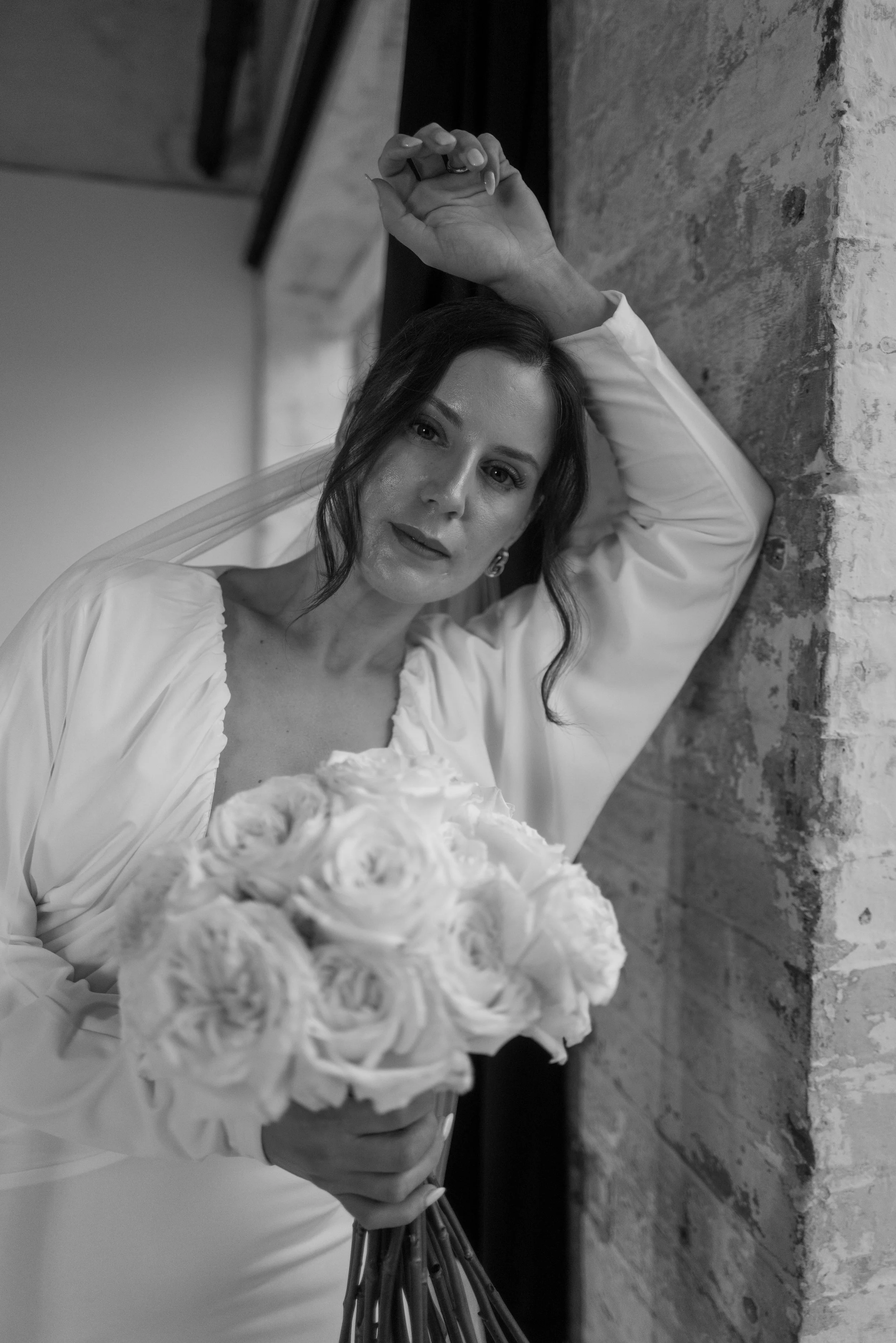 Black and white photo of a woman holding a bouquet of roses, leaning against a brick wall, with her arm raised and resting on her head.