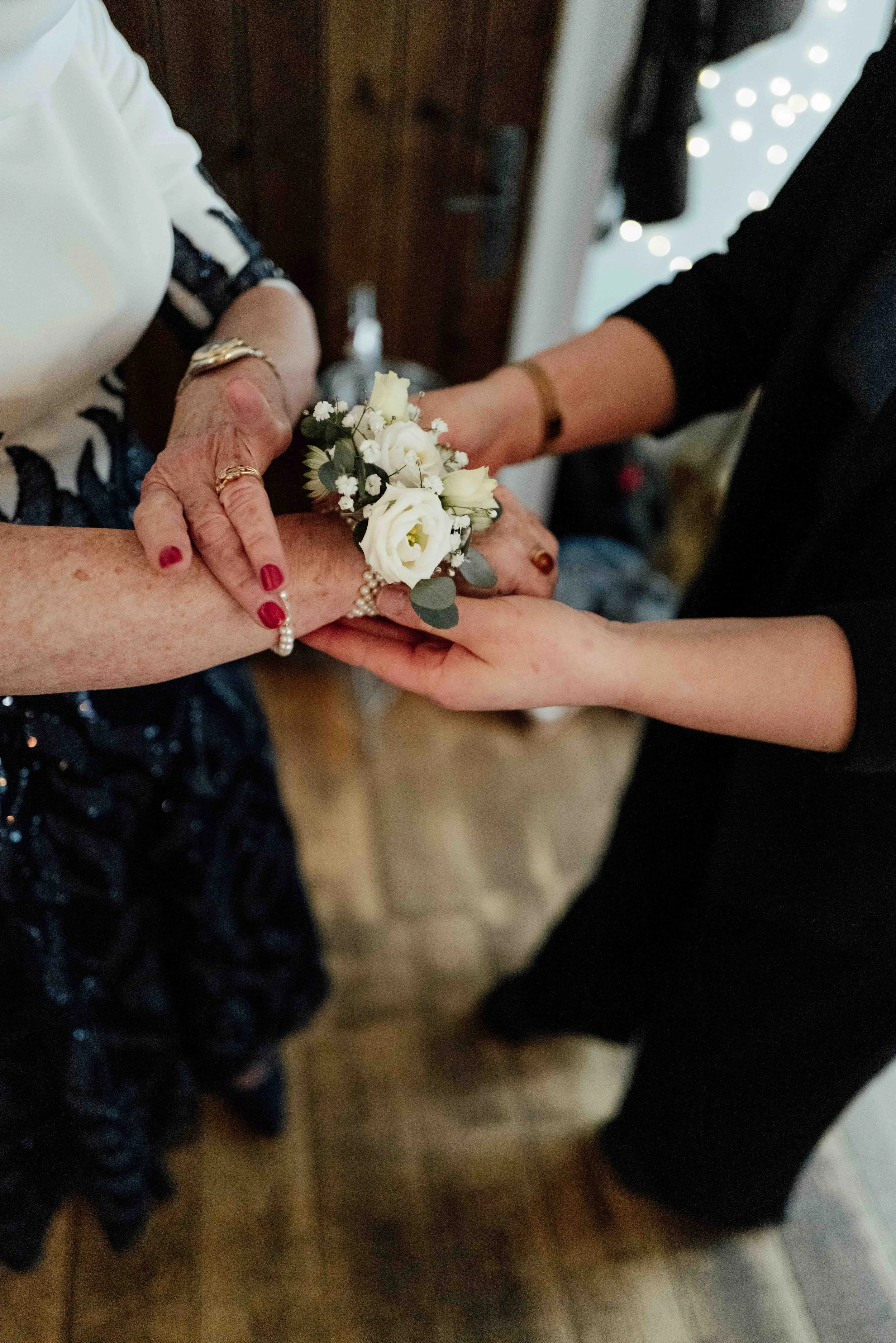 Two women are holding hands, with one woman presenting a small white flower corsage and the other holding her hand. The women are dressed in formal attire, likely at a special event or ceremony.