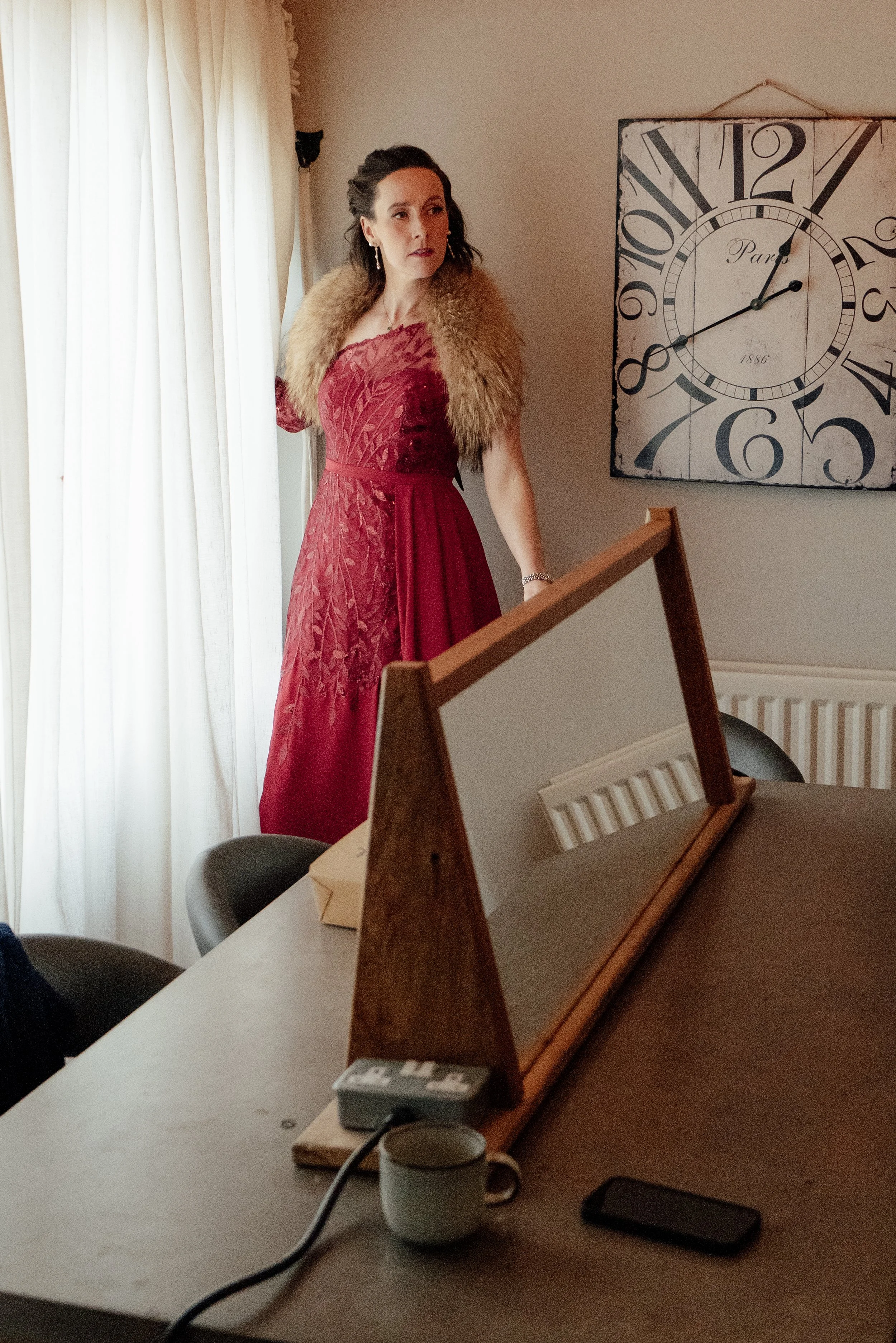 A woman in a red dress with a fur stole standing near a window in a dining room, with a large wall clock and a mirror on the table.