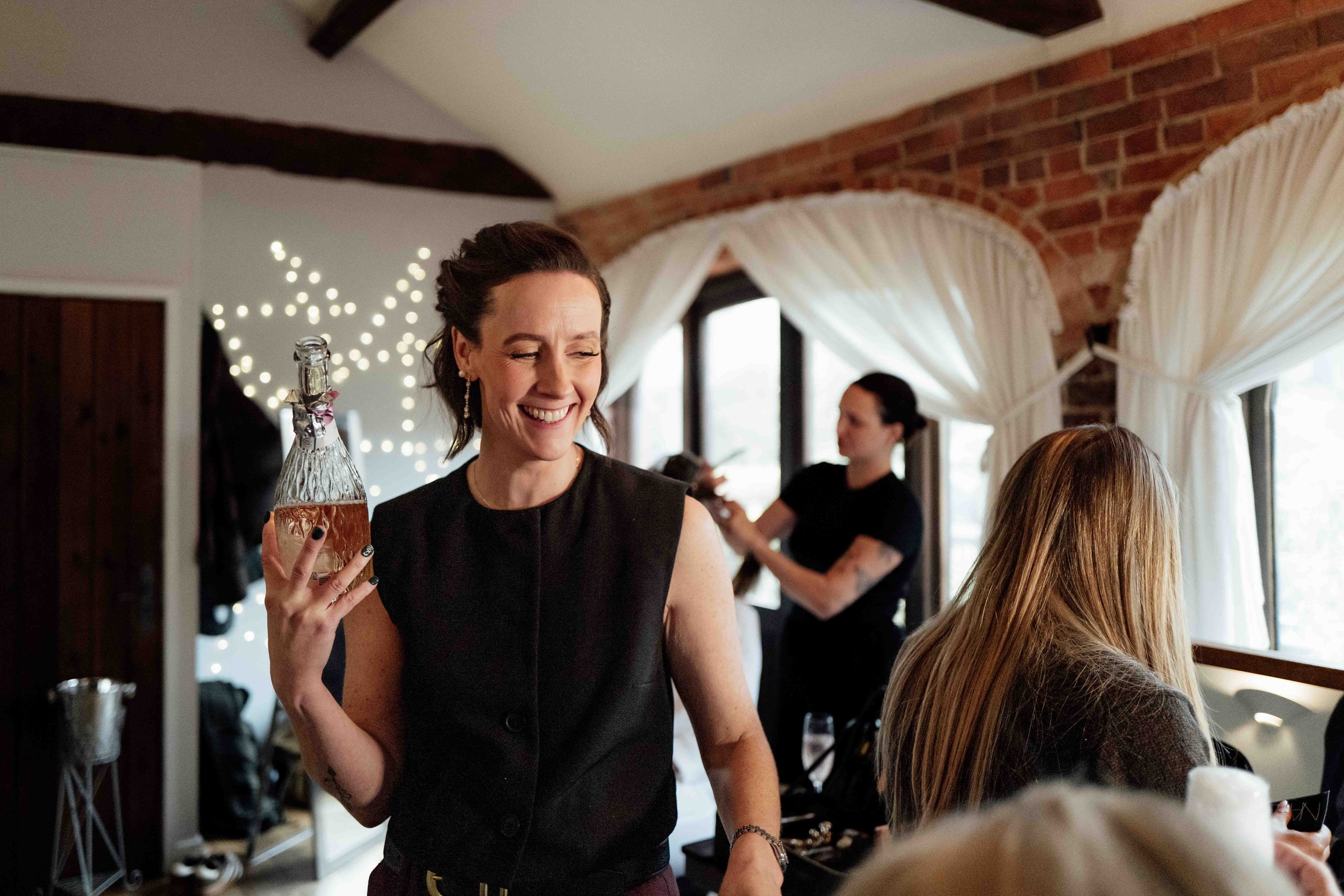 A woman smiling and holding a bottle of rosé wine inside a room with brick walls and curtains, with another woman in the background styling hair.