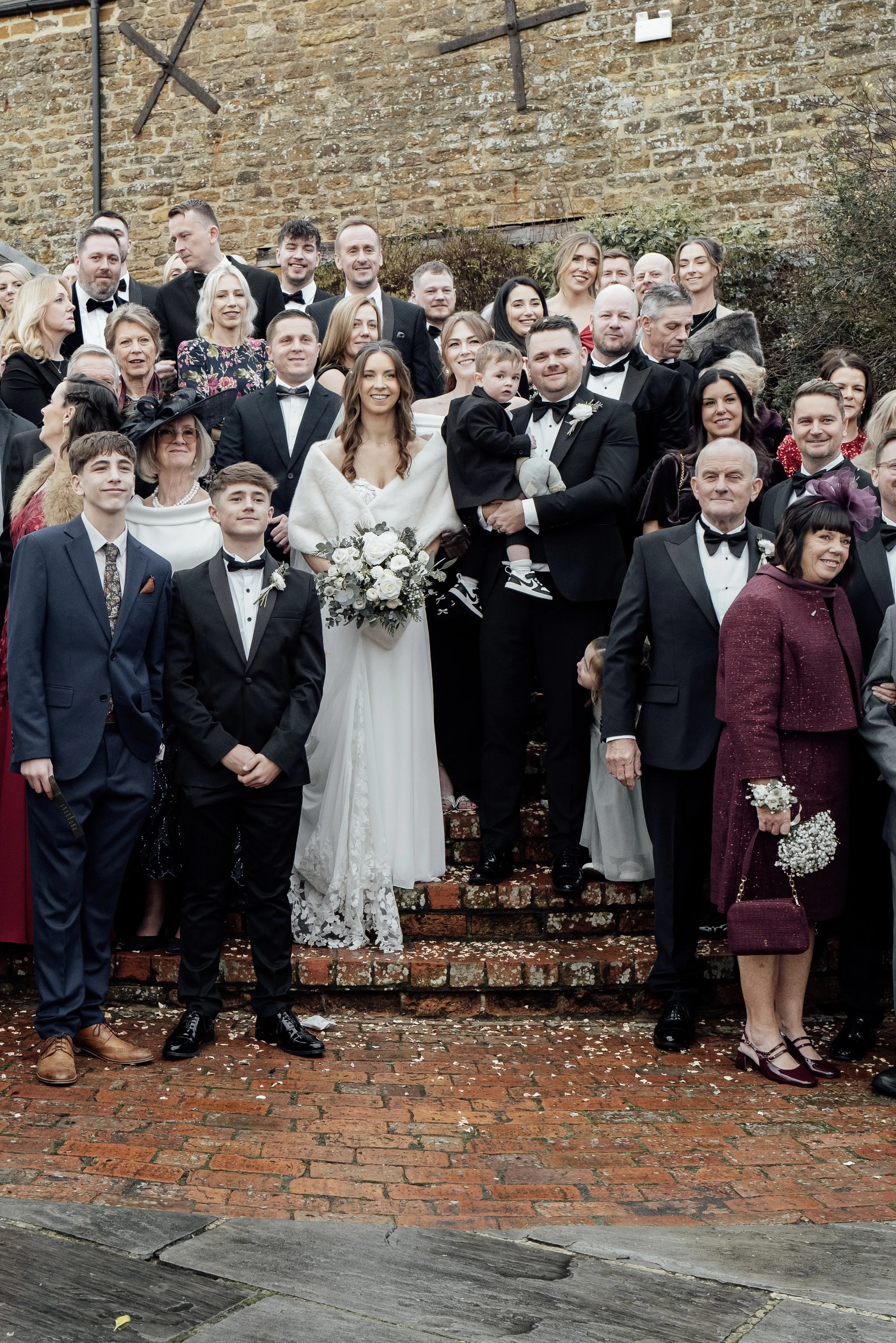 Group of wedding guests, including the bride and groom, posing outdoors on brick steps with a stone wall background, during daytime.