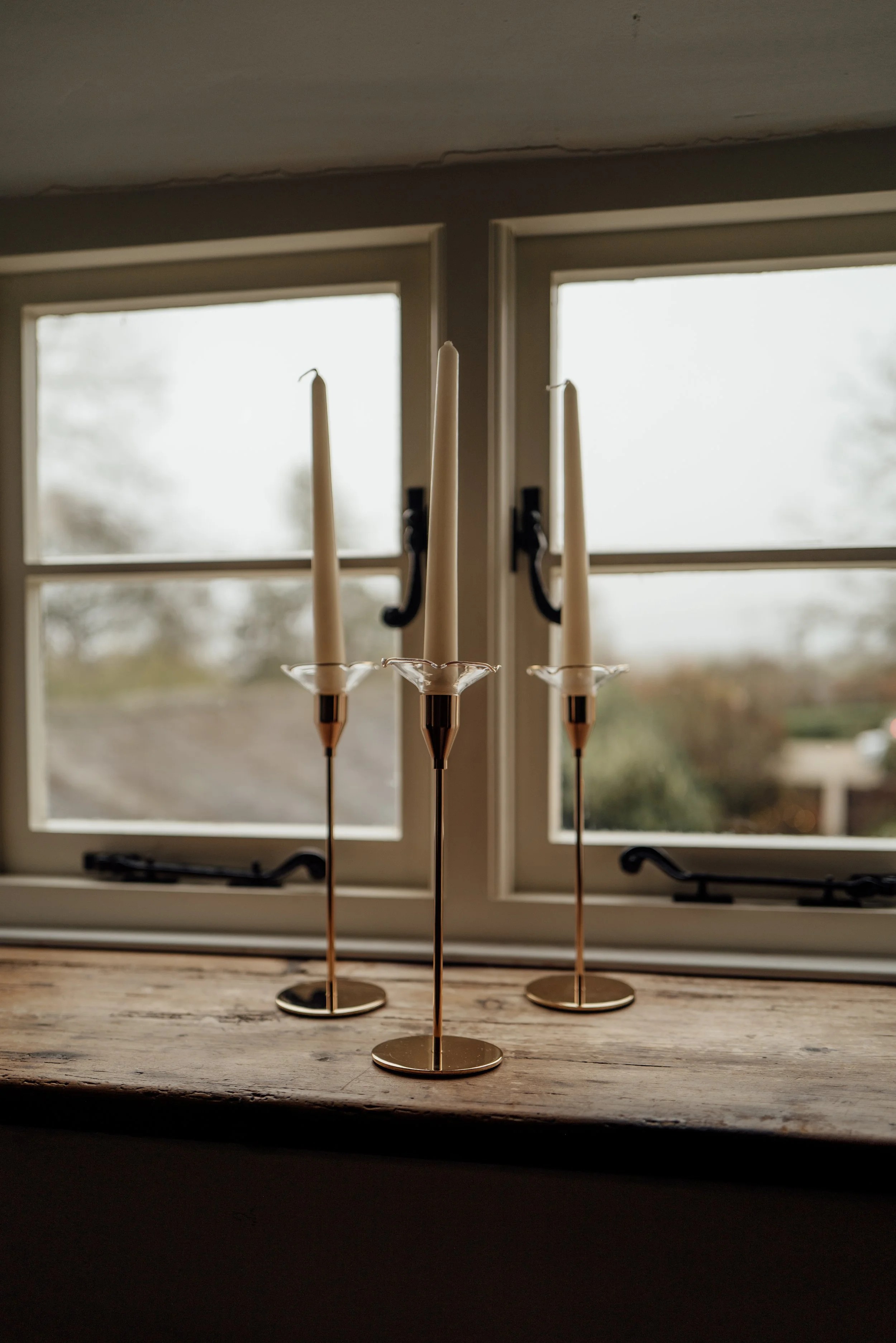 Three brass candle holders with unlit white candles on a wooden windowsill, with a window showing an outdoor view of trees and sky.