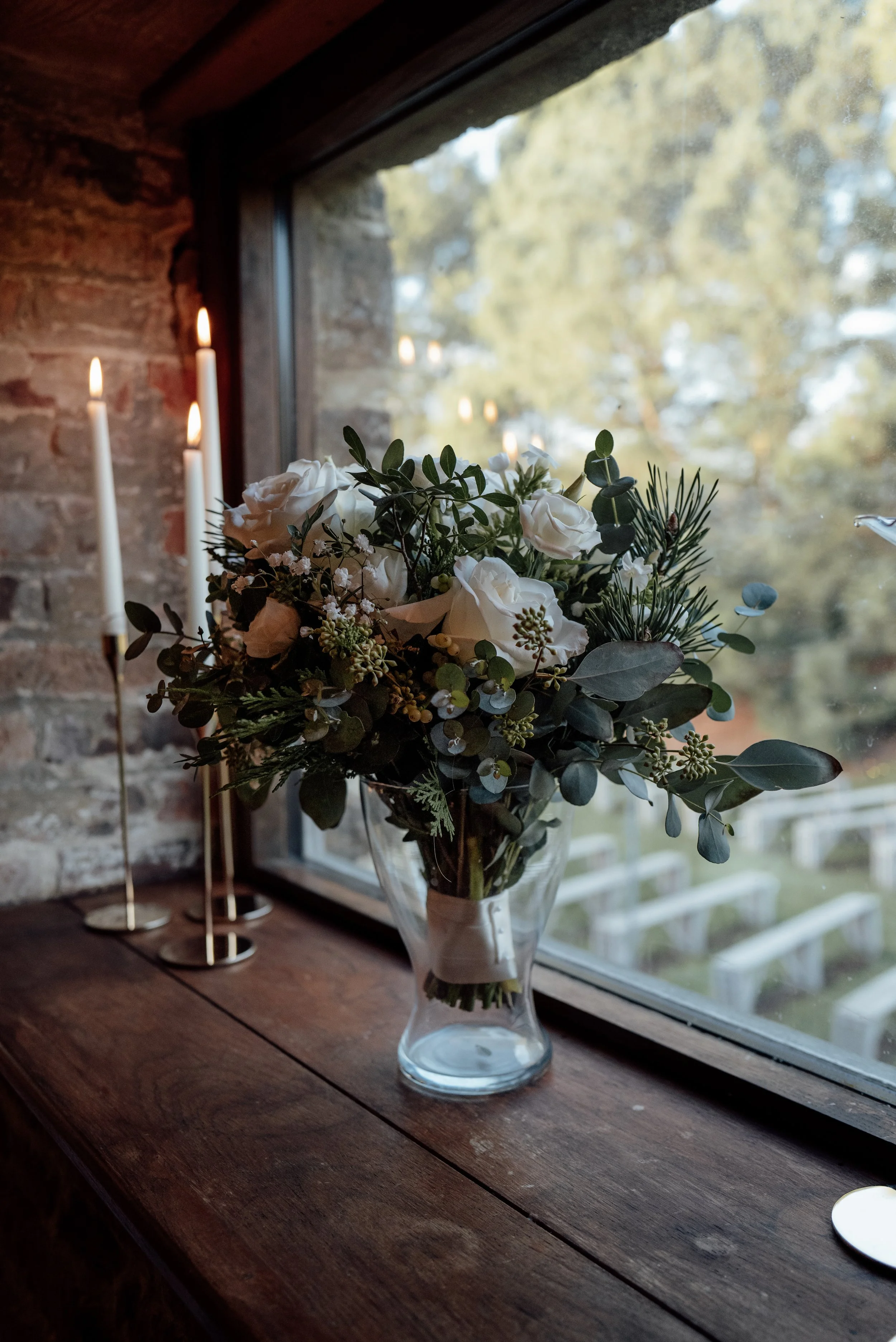 A glass vase with white roses, eucalyptus, and greenery on a wooden windowsill with candles in the background.
