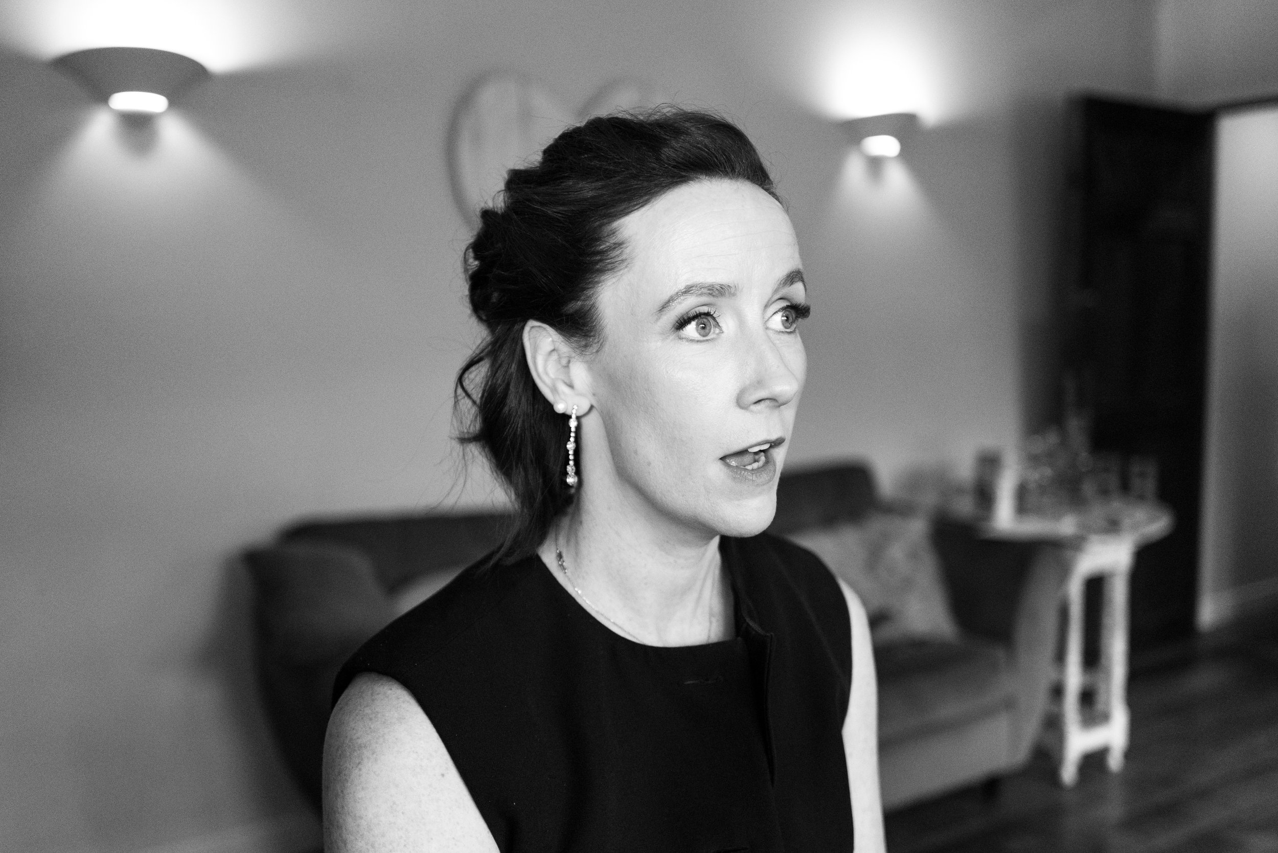 Black and white photo of a woman with dark hair styled in an updo, wearing earrings and a sleeveless top, indoors with blurred background including a sofa and a side table.