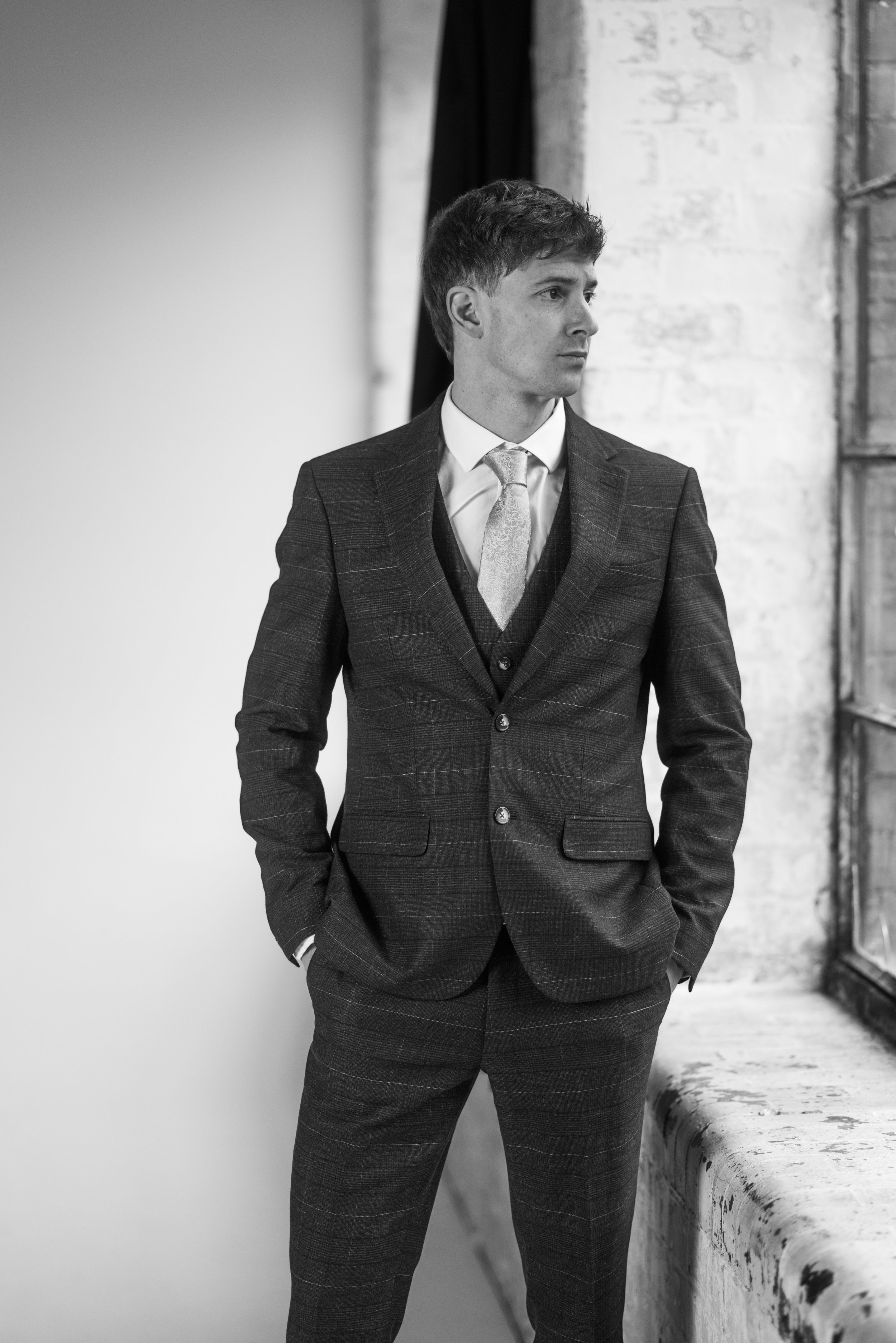 A man in a dark, checked suit with a light tie, standing indoors near a window, looking to the side with hands in pockets.
