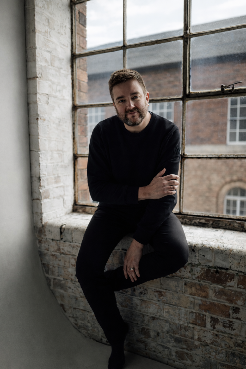 A man with a beard and short hair, wearing a black shirt and pants, sitting on a window ledge in front of a large industrial-style window with a brick exterior visible outside.