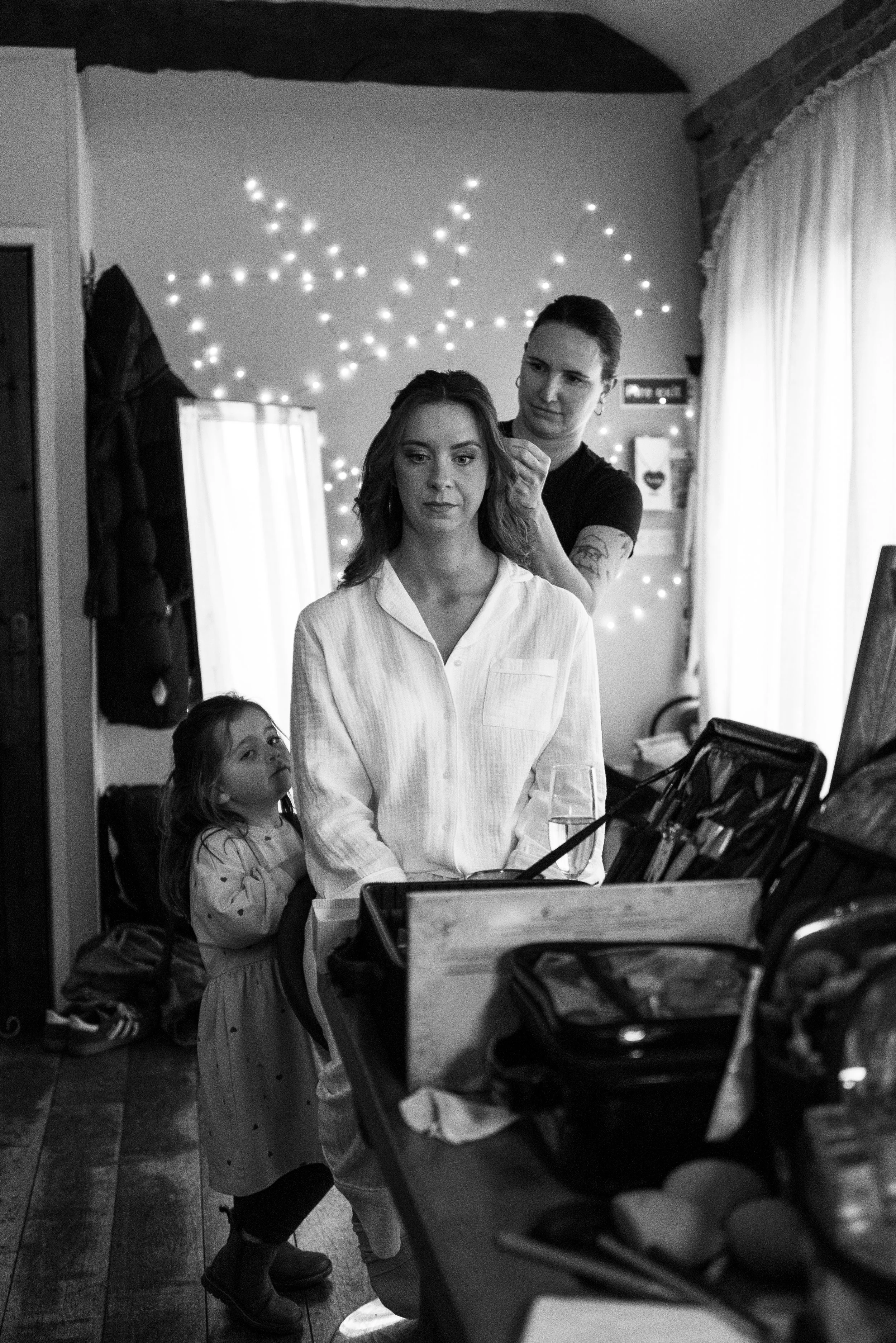 A woman sitting at a desk with makeup tools, while another woman styles her hair behind a mirror. A young girl stands nearby watching. The scene is illuminated by string lights on the wall.