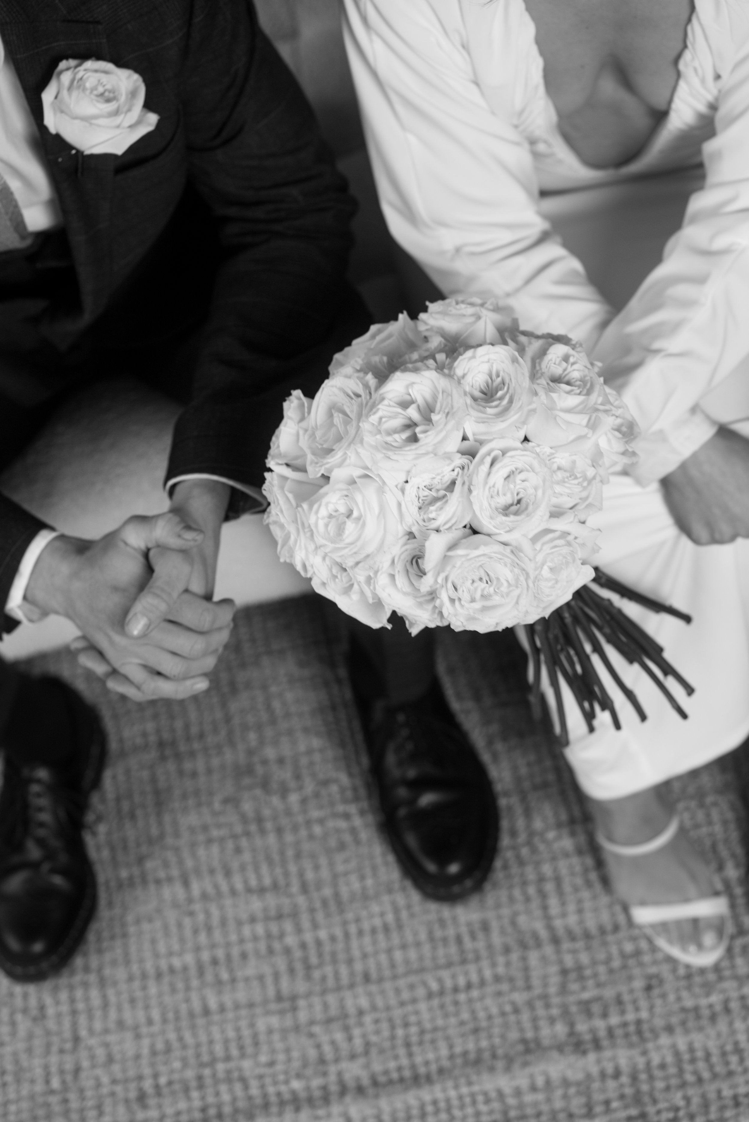A black and white photo of two people holding hands, one wearing a suit with a rose on his lapel, and the other in a white dress holding a bouquet of roses.