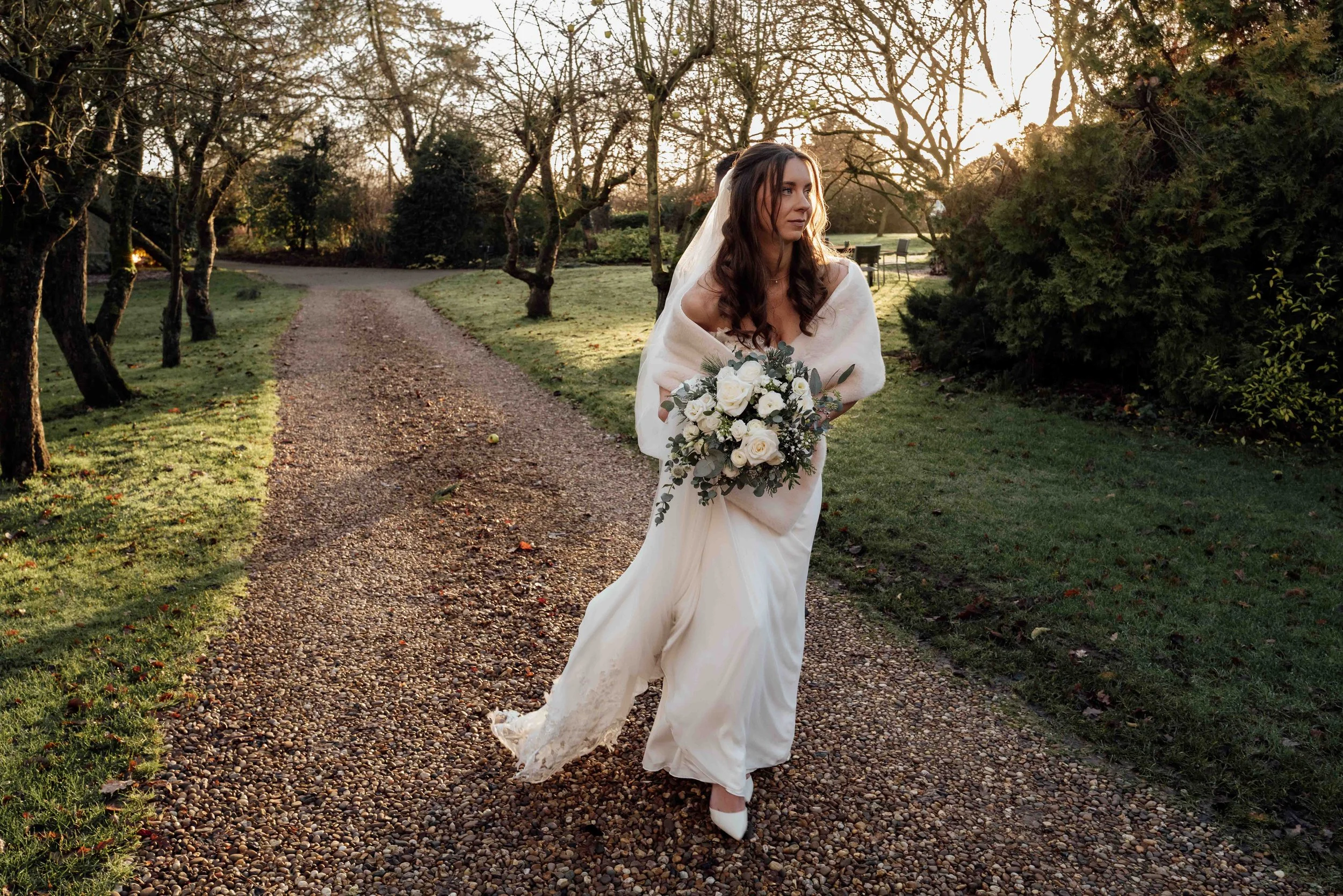 A woman in a wedding dress holding a bouquet of white flowers walking on a gravel path in a park at sunset.