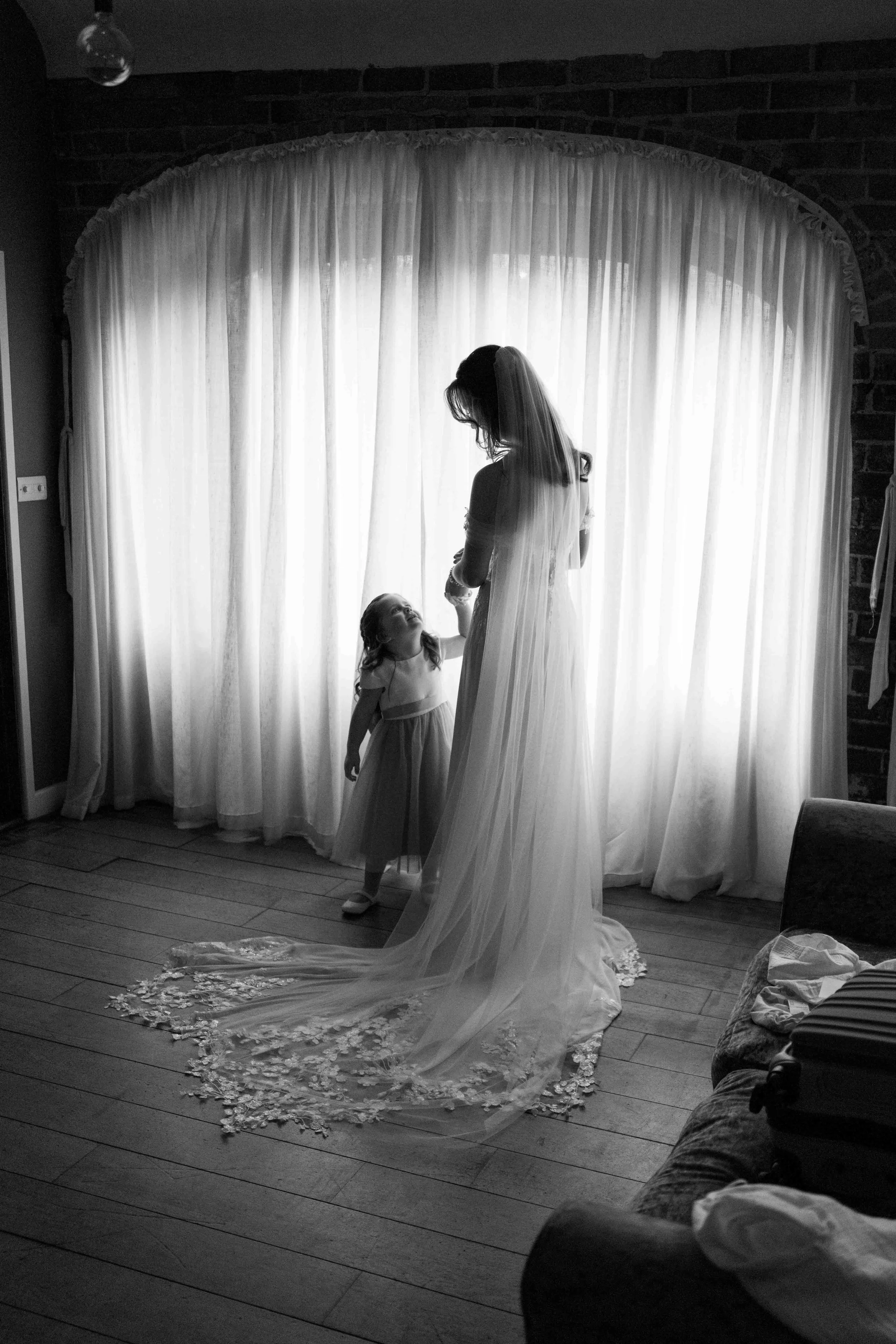 A bride in a wedding dress with a long veil stands in front of a window with sheer curtains, holding hands with a young girl in a dress, inside a room with wooden flooring and brick wall.