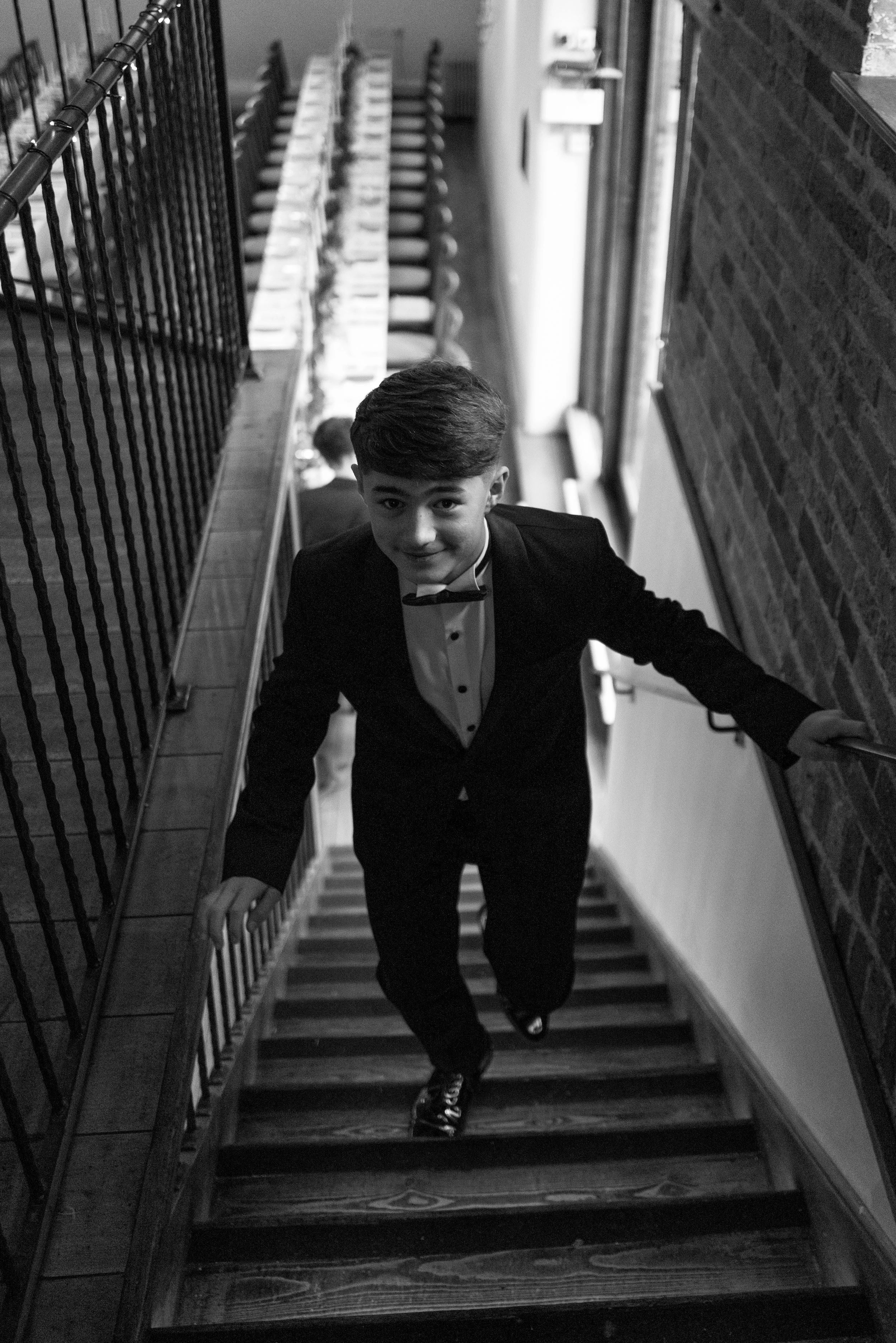 A young man in a tuxedo and bow tie climbing a staircase inside a building, smiling at the camera.