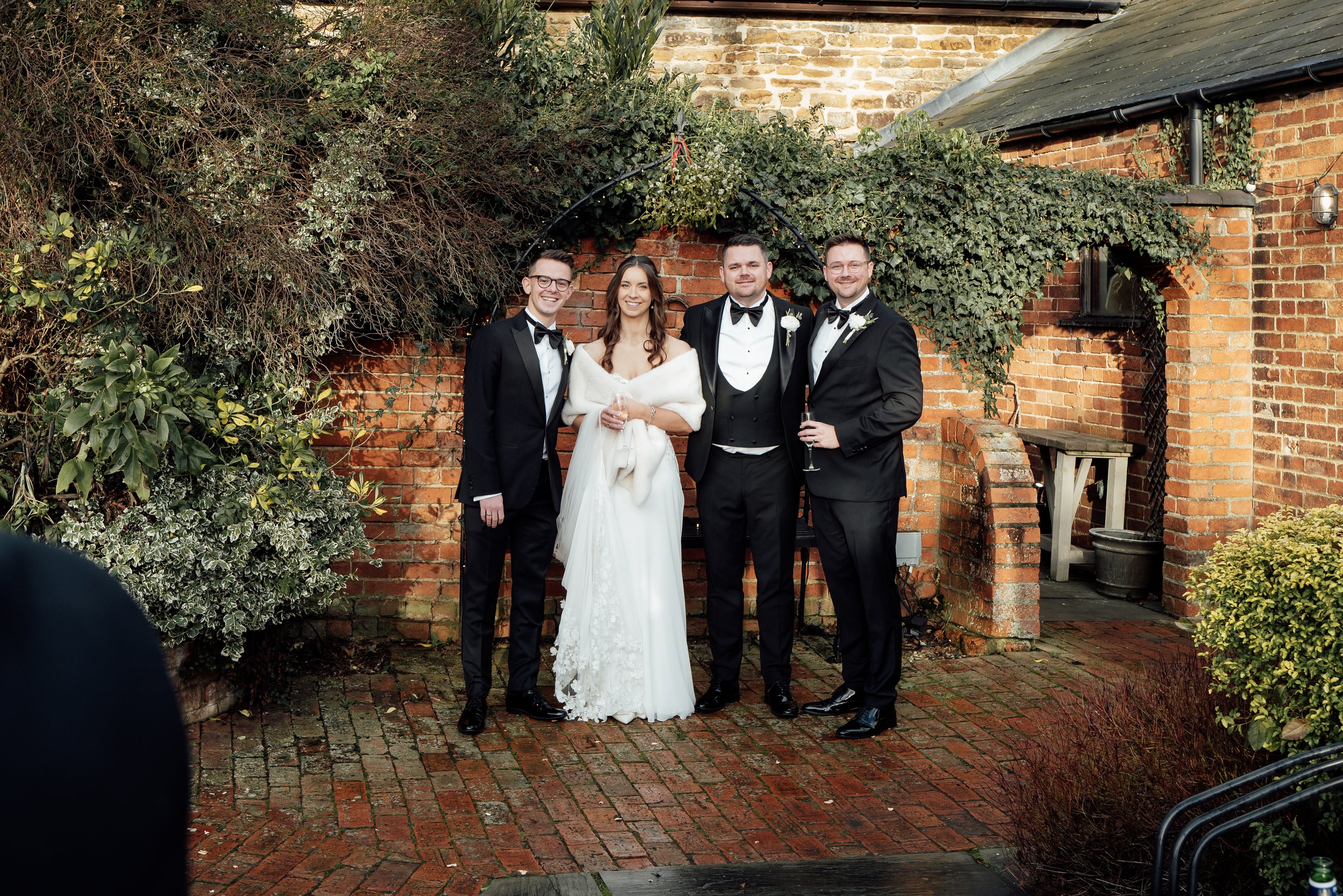A group of four people in formal wedding attire standing outdoors in front of a brick wall with greenery, smiling at the camera. The bride wears a white dress and holds a glass, and the three men are in black tuxedos with bow ties, holding glasses of